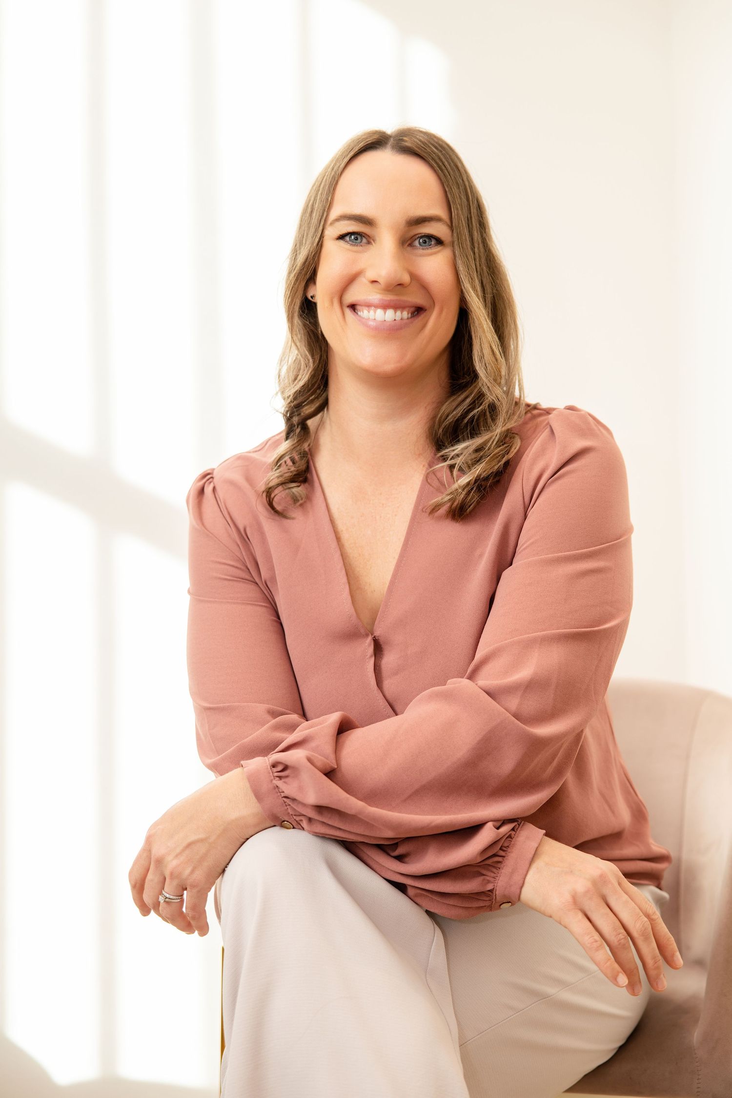 Smiling woman in pink blouse sitting relaxed in bright white room, professional portrait photo.