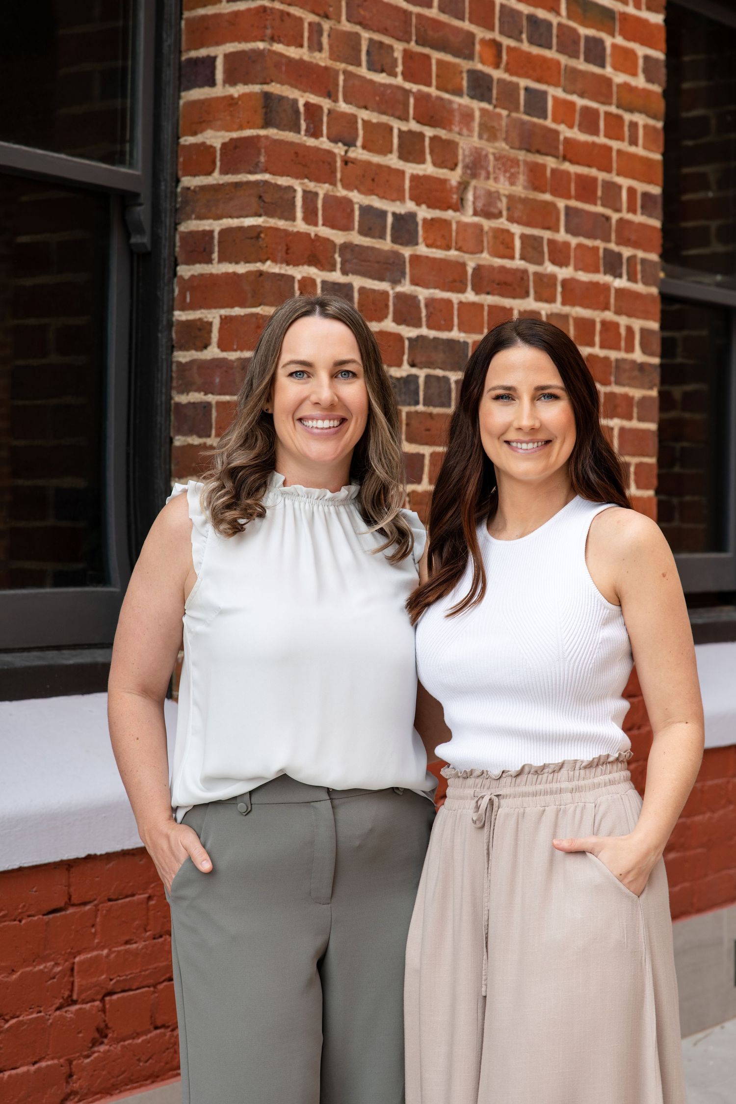 Two smiling women in neutral tops standing together in front of a red brick wall.