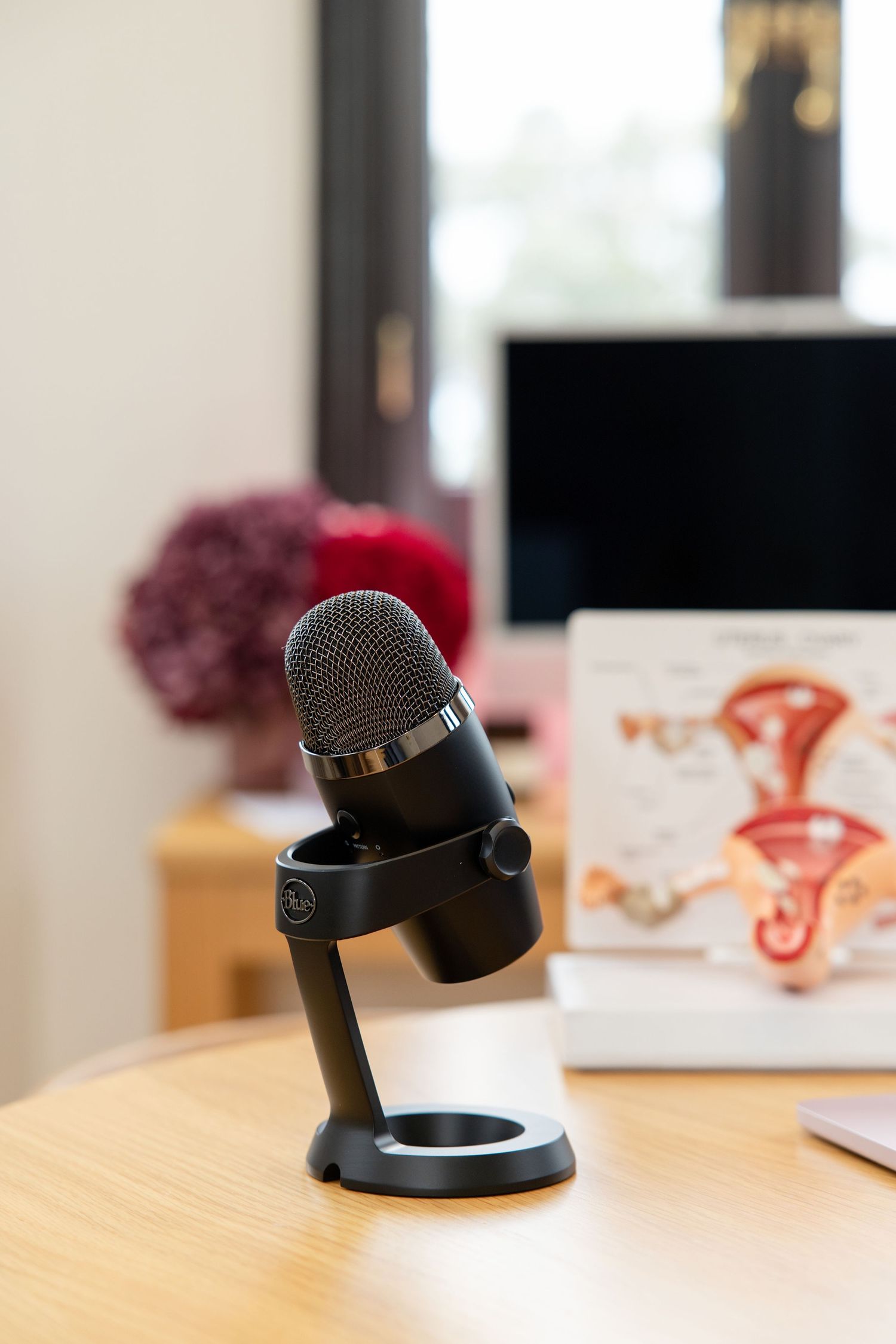 Black USB microphone on a wooden desk with anatomical diagram and computer monitor in background.
