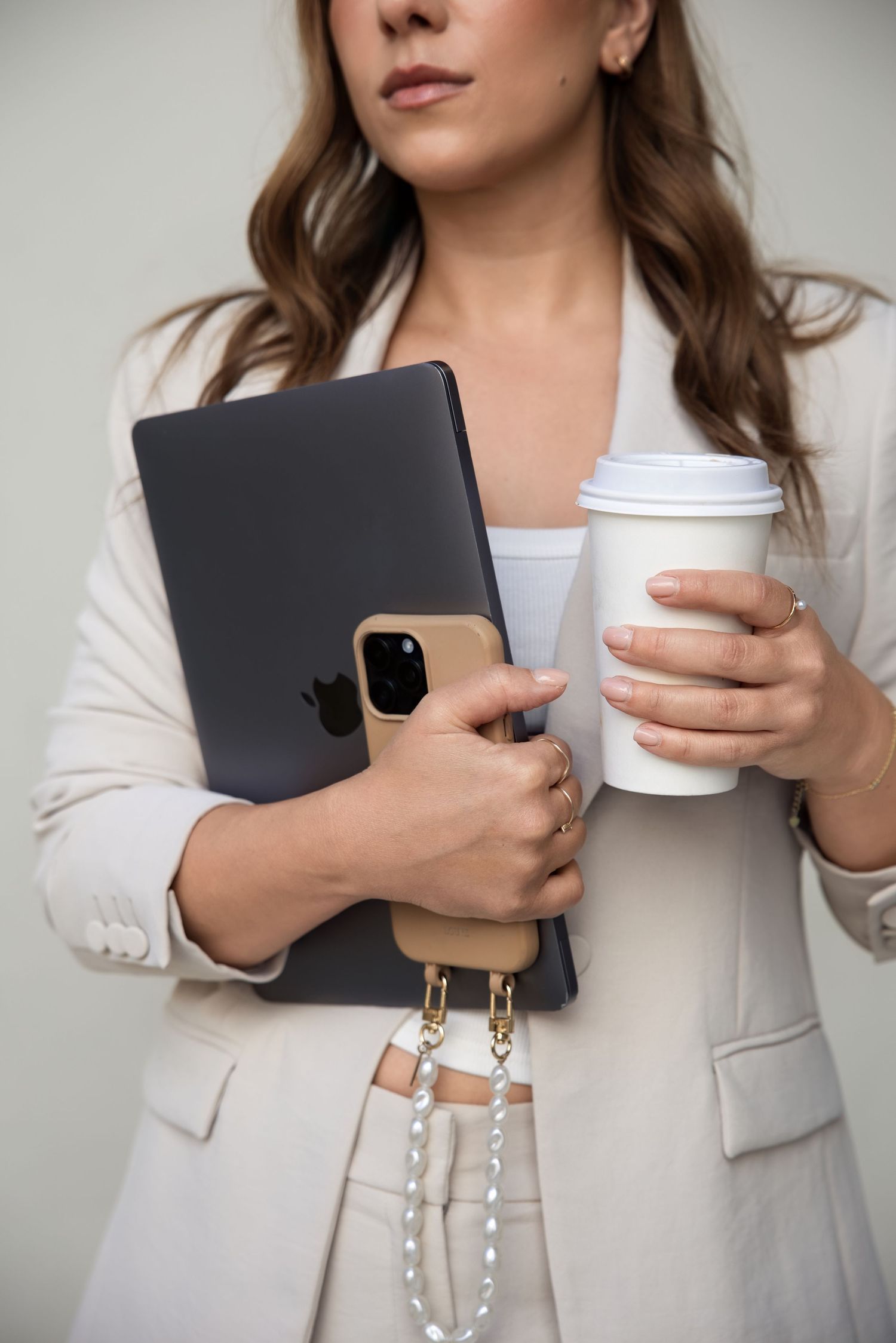 Woman in white blazer holding Apple iPad, iPhone with beige case, and white takeaway coffee cup.