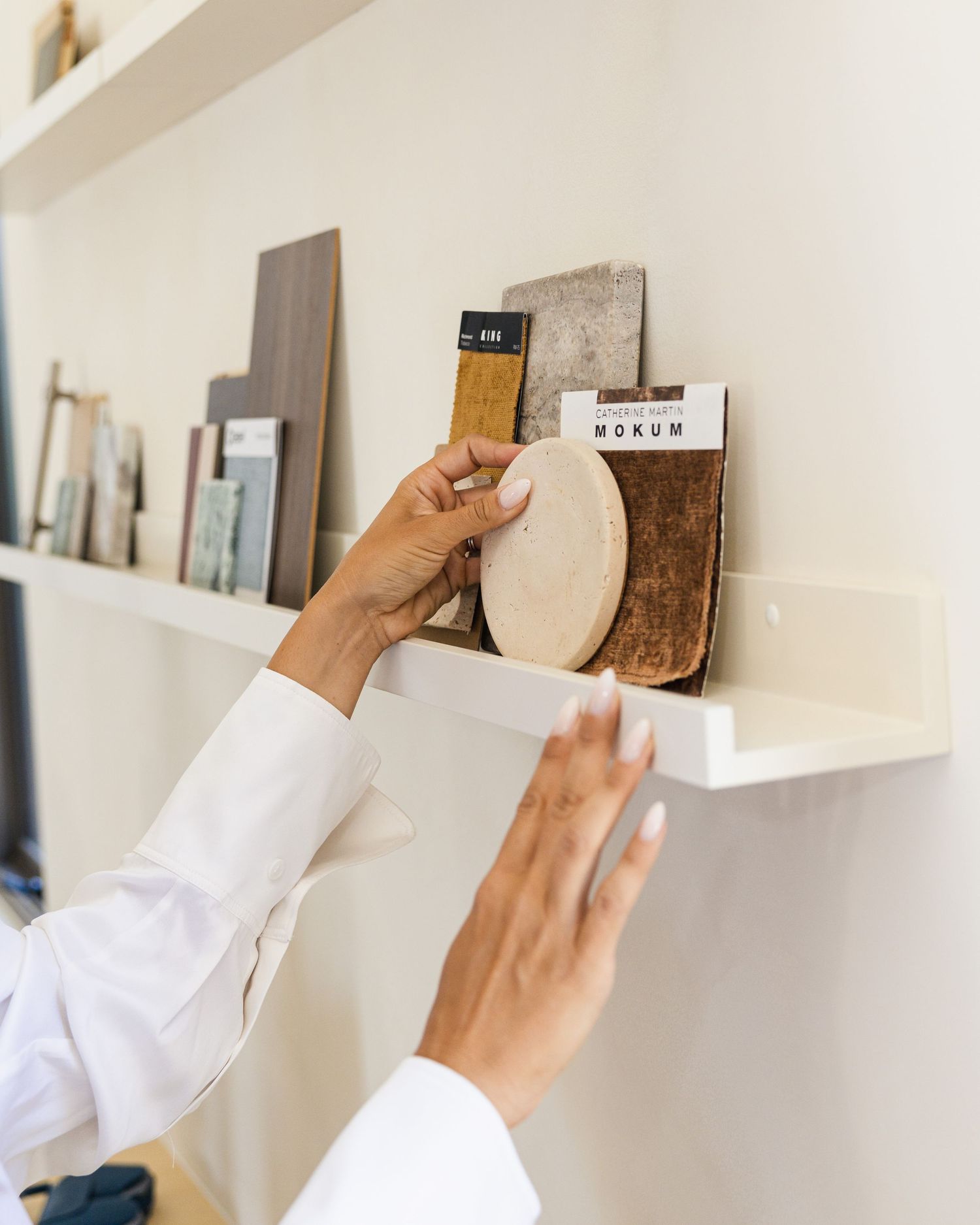 Person in white blazer arranging material samples on a minimalist display shelf in a design studio.