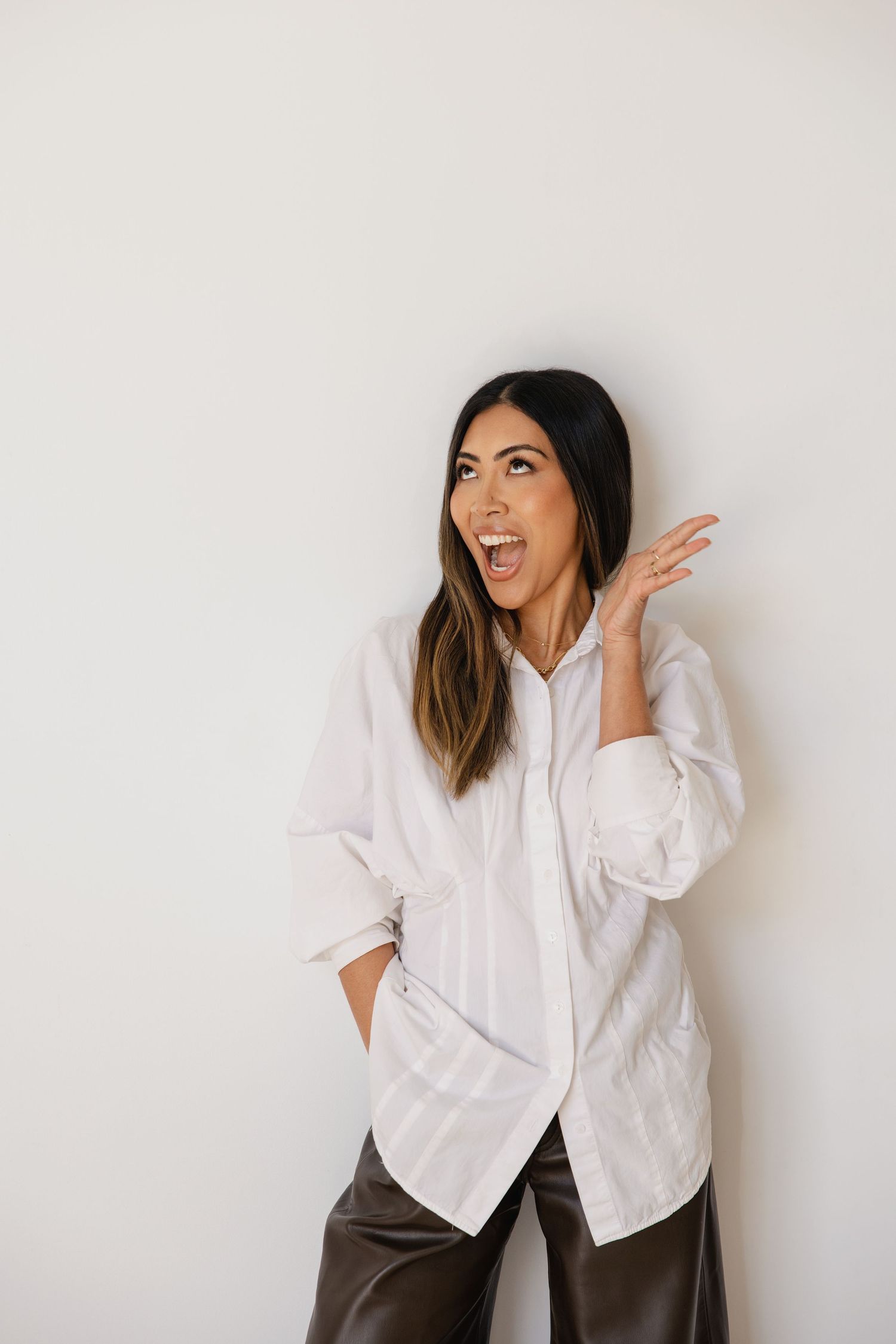 Woman in white oversized shirt and dark pants poses expressively against a light background.
