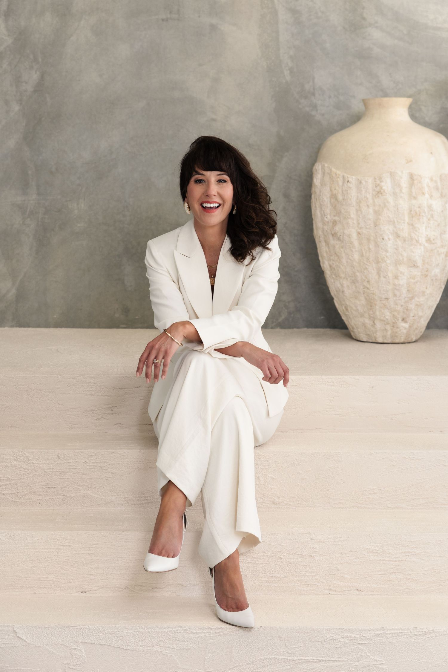 A person in an elegant white suit sits casually on a light-colored floor next to a large decorative cream vase.