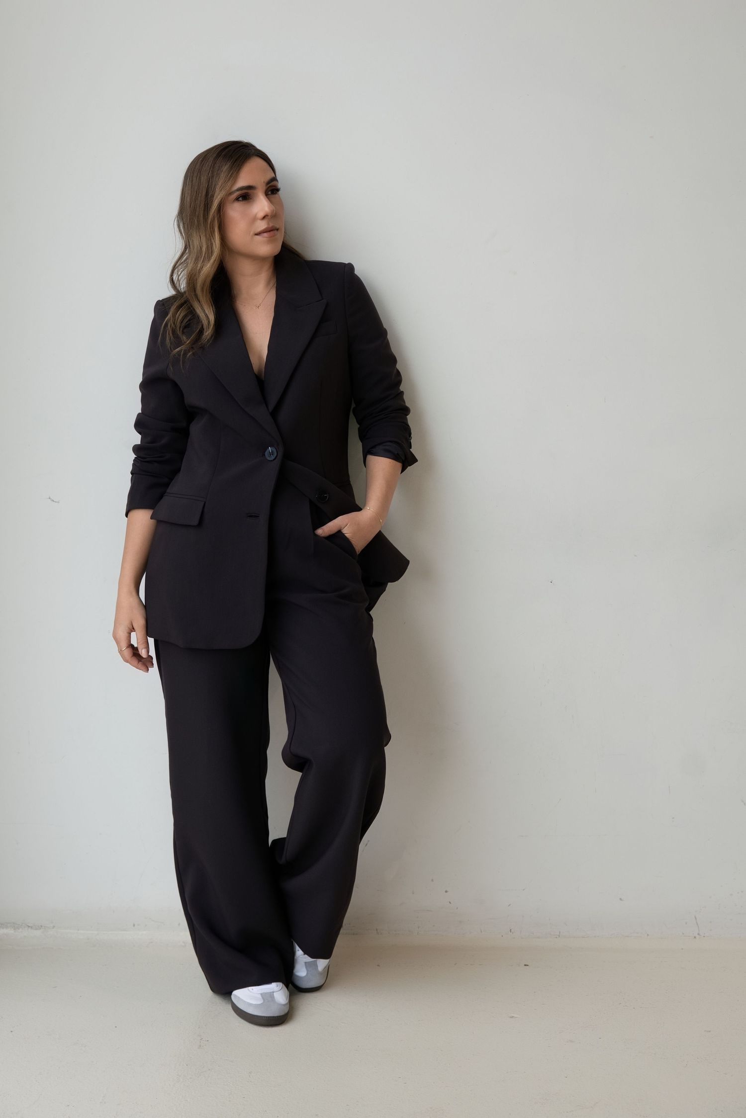 Stylish woman in black blazer and wide-leg trousers leaning against white wall in casual power pose.