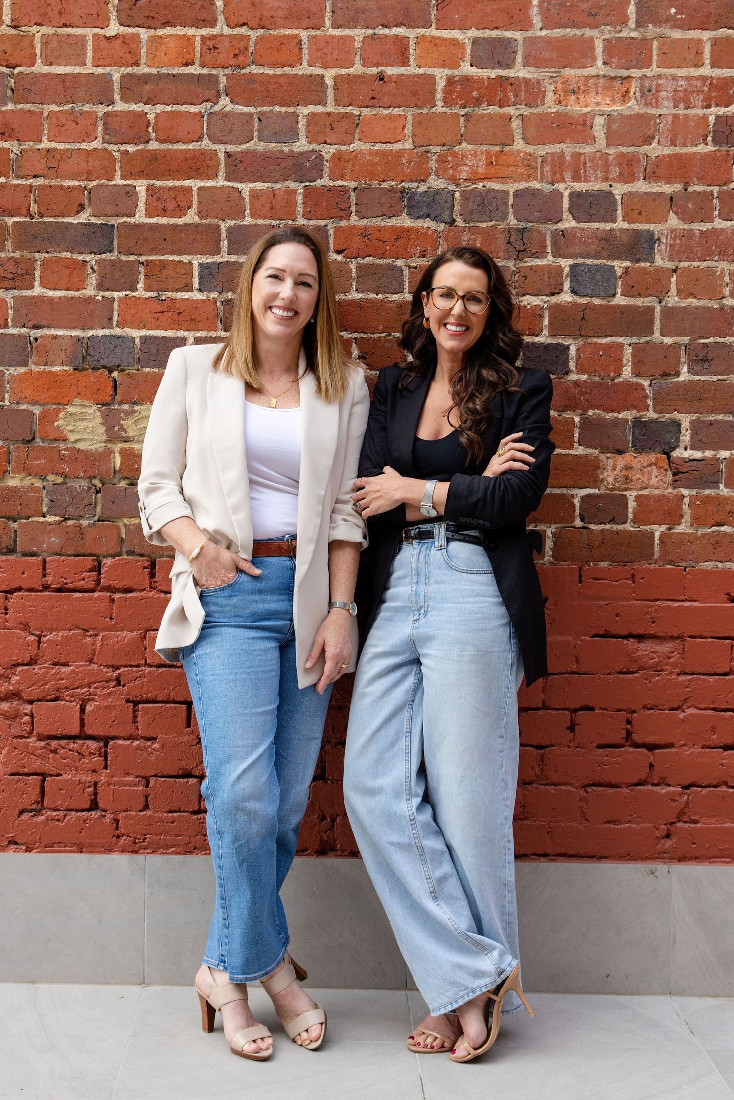 Two smiling women in blazers and jeans posing against a rustic red brick wall.