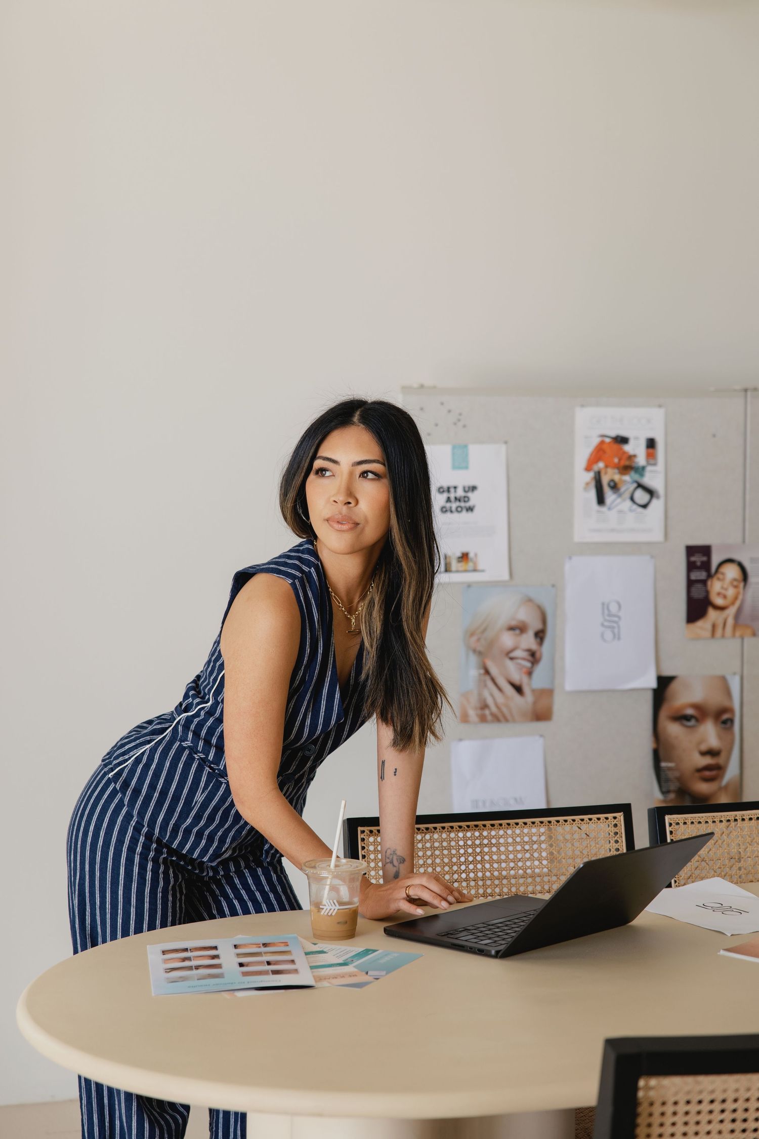 A woman in a striped jumpsuit leans on a round table with a laptop in a modern workspace with a mood board.
