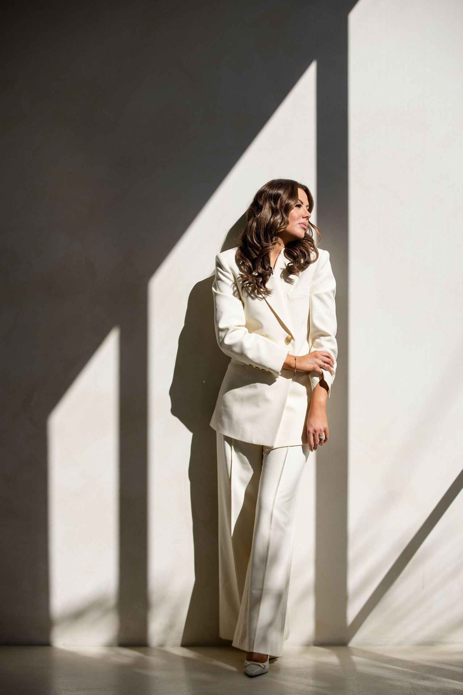 Stylish woman in white blazer and wide-leg trousers posing in sunlit room with geometric shadows.