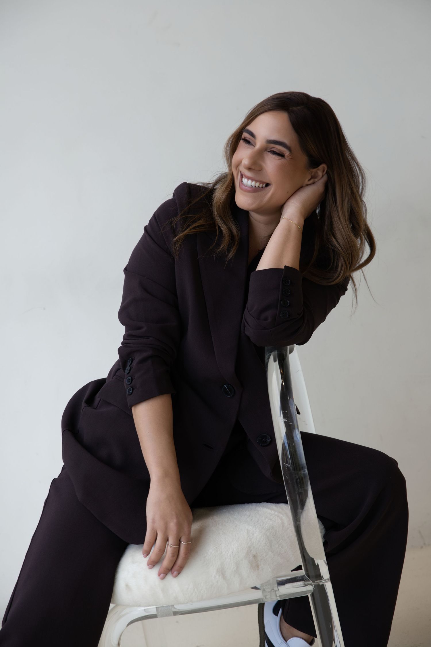 Smiling woman in black blazer suit sitting on chair against white background.