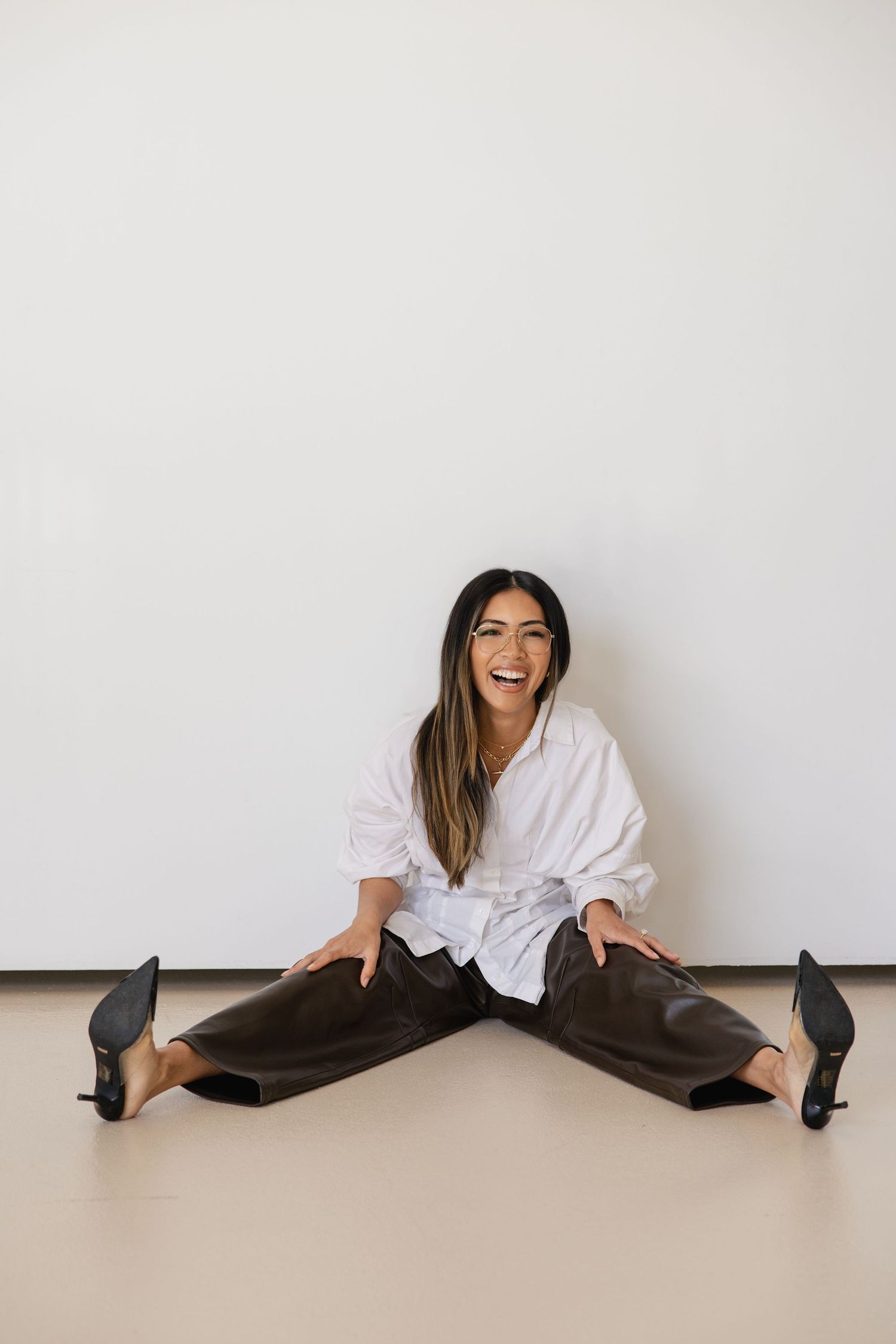 Smiling woman in white shirt and dark leather pants doing splits on floor against white wall.