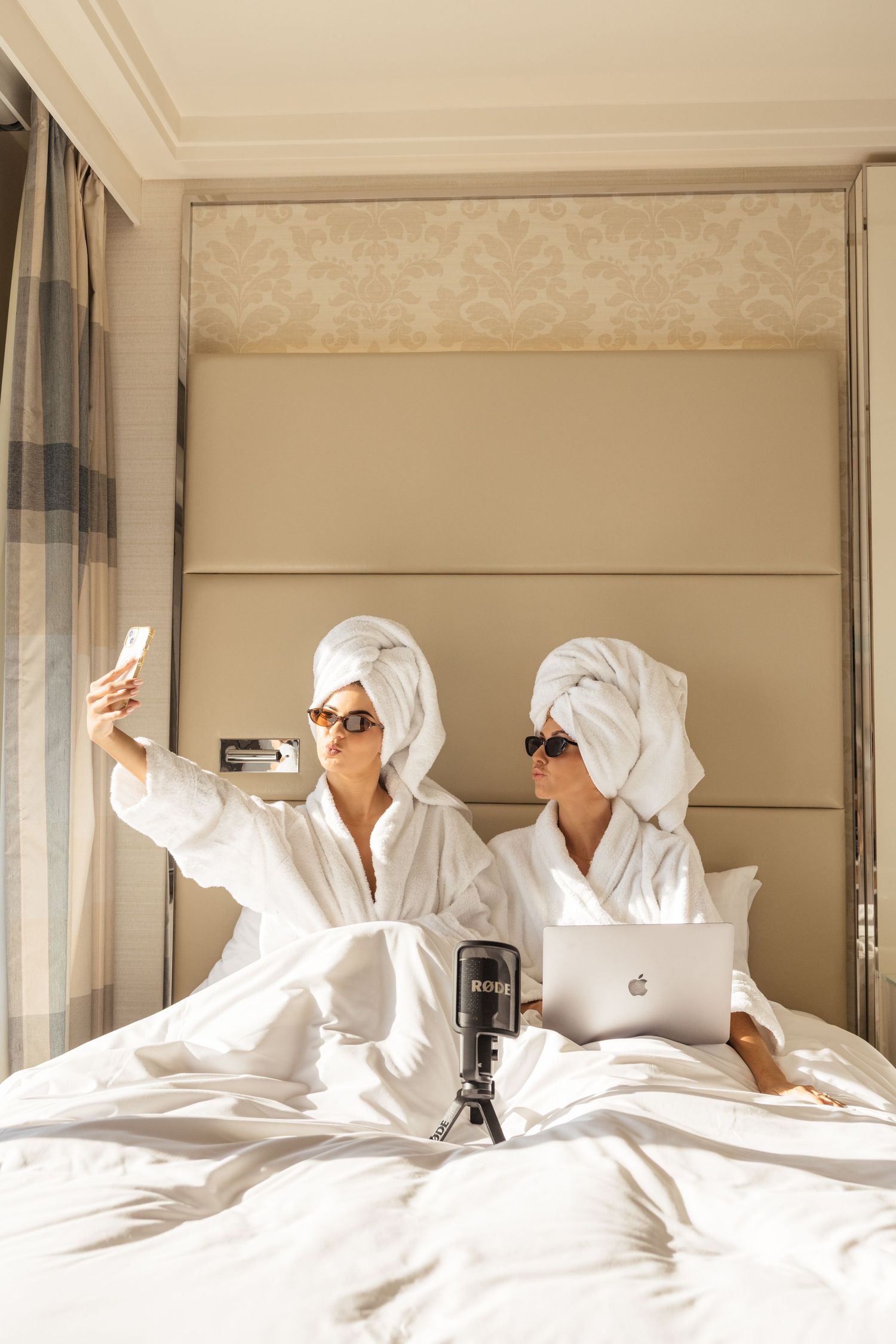 Group of people relaxing in white bathrobes and towels on a large hotel bed while using phones and devices.