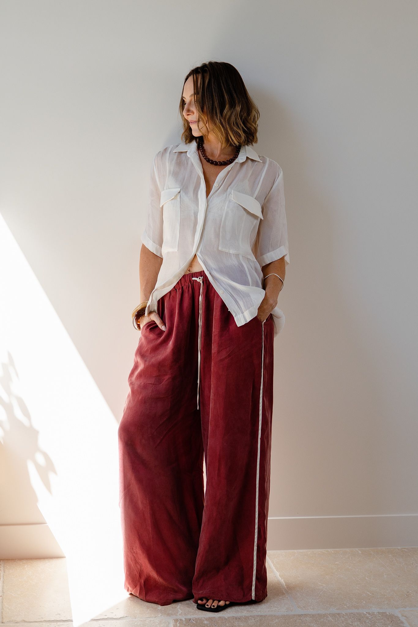 Woman wearing wide-leg burgundy velvet pants with white stripe and sheer white button-up shirt, posing against light wall.