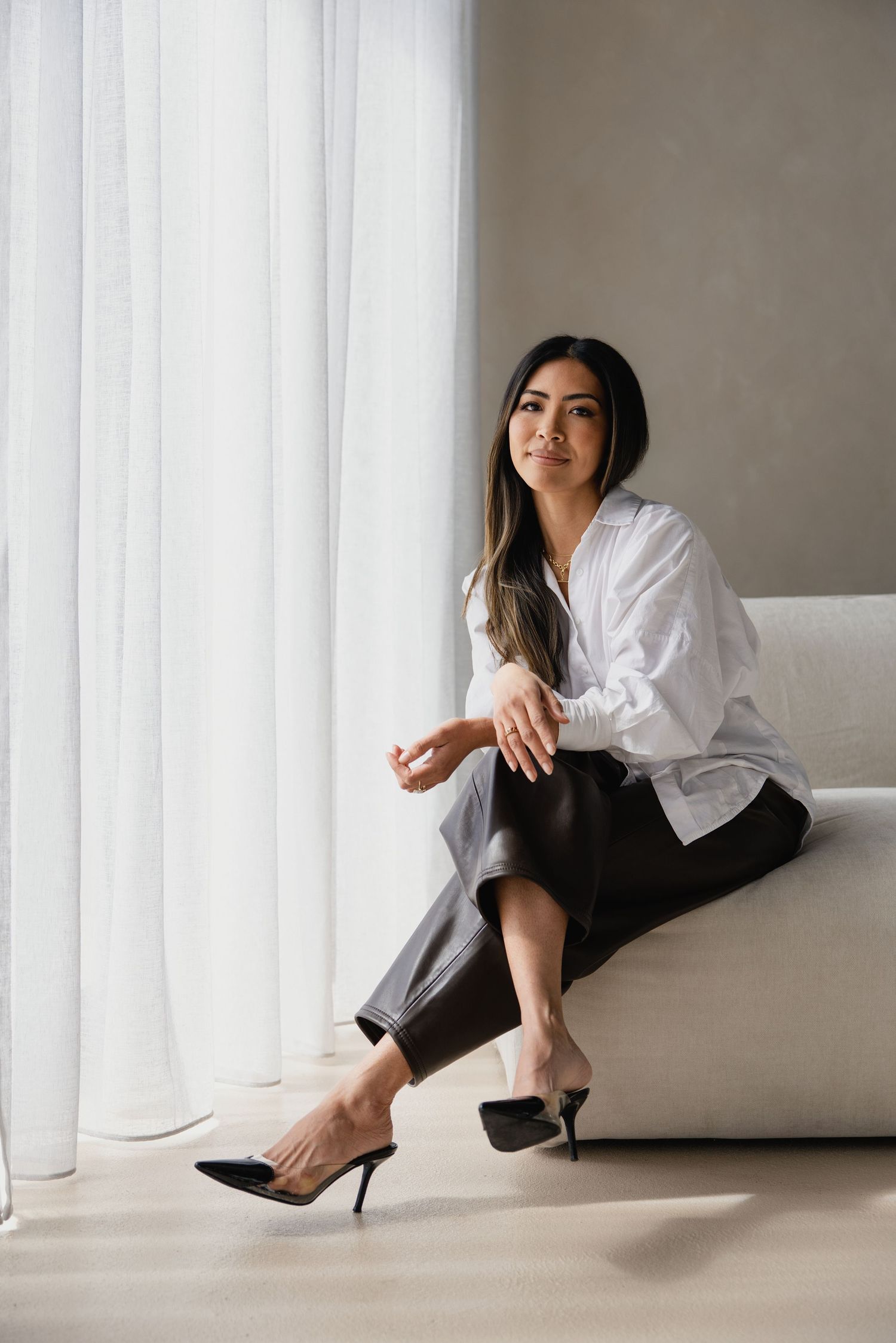 Woman in white blouse and black leather pants sitting on sofa near sheer curtains wearing black heeled mules.