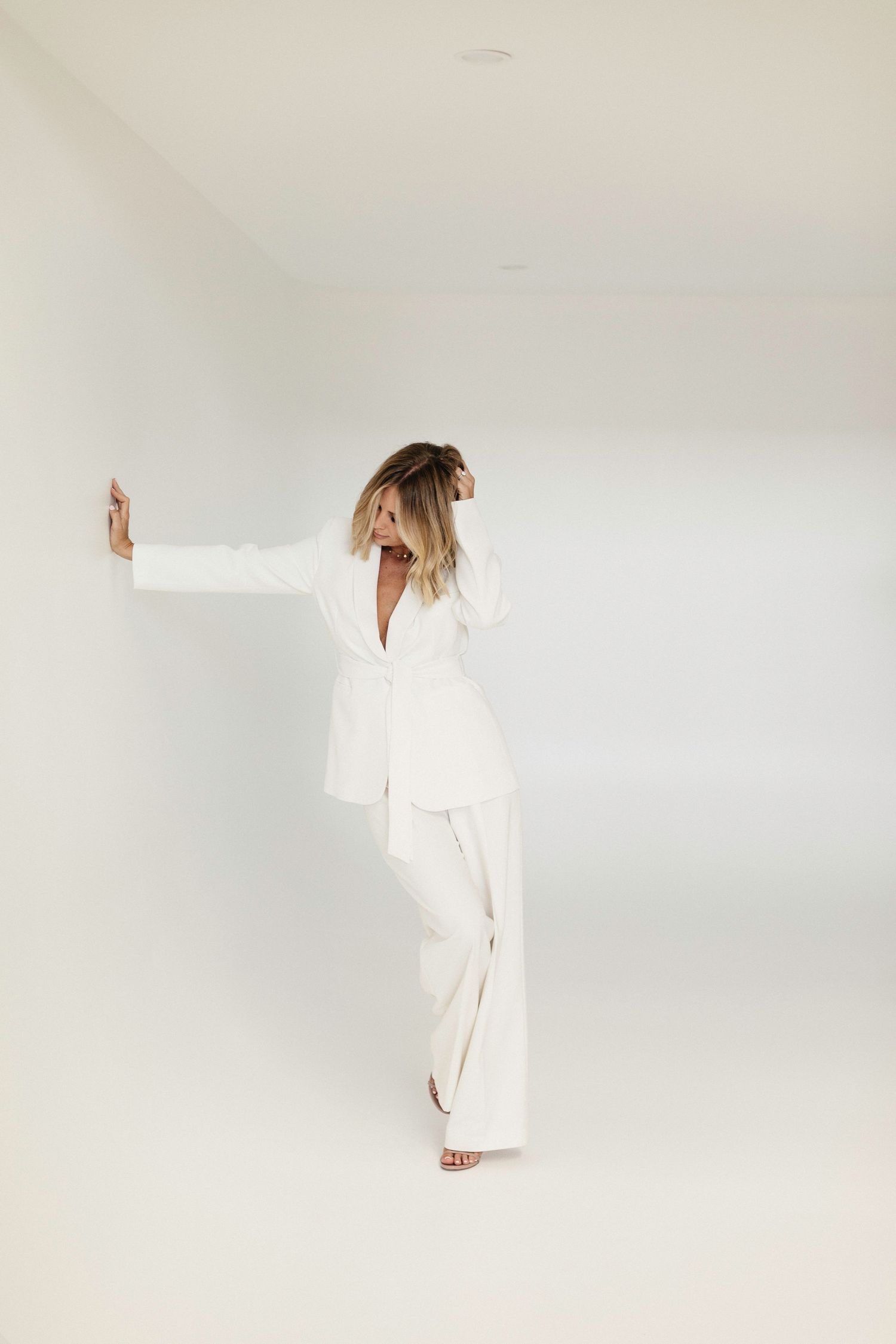 Woman in white blazer and wide-leg pants posing against a white wall in a minimalist studio setting.