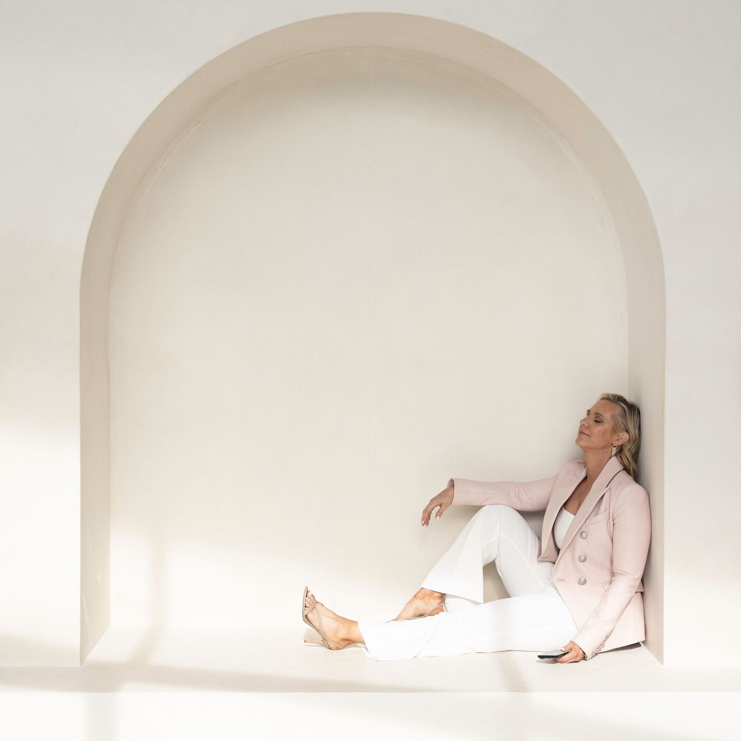 Woman in pink blazer and white pants sitting relaxed inside a minimalist arched alcove with soft neutral tones.