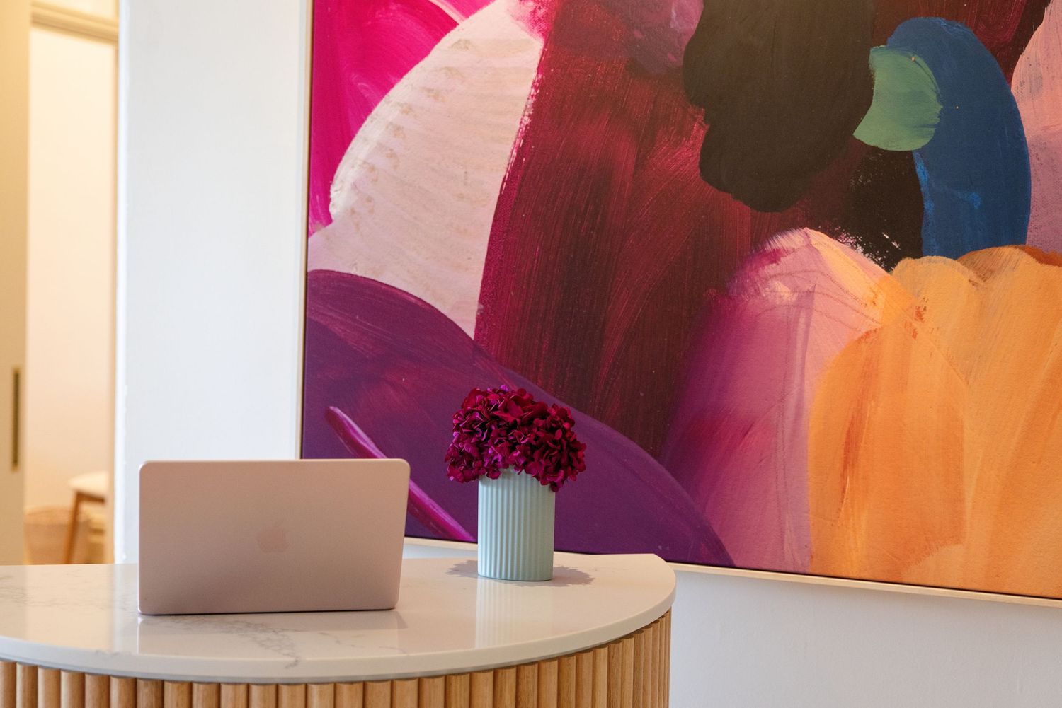 Modern reception desk with laptop and pink flowers in front of a vibrant colorful abstract painting.