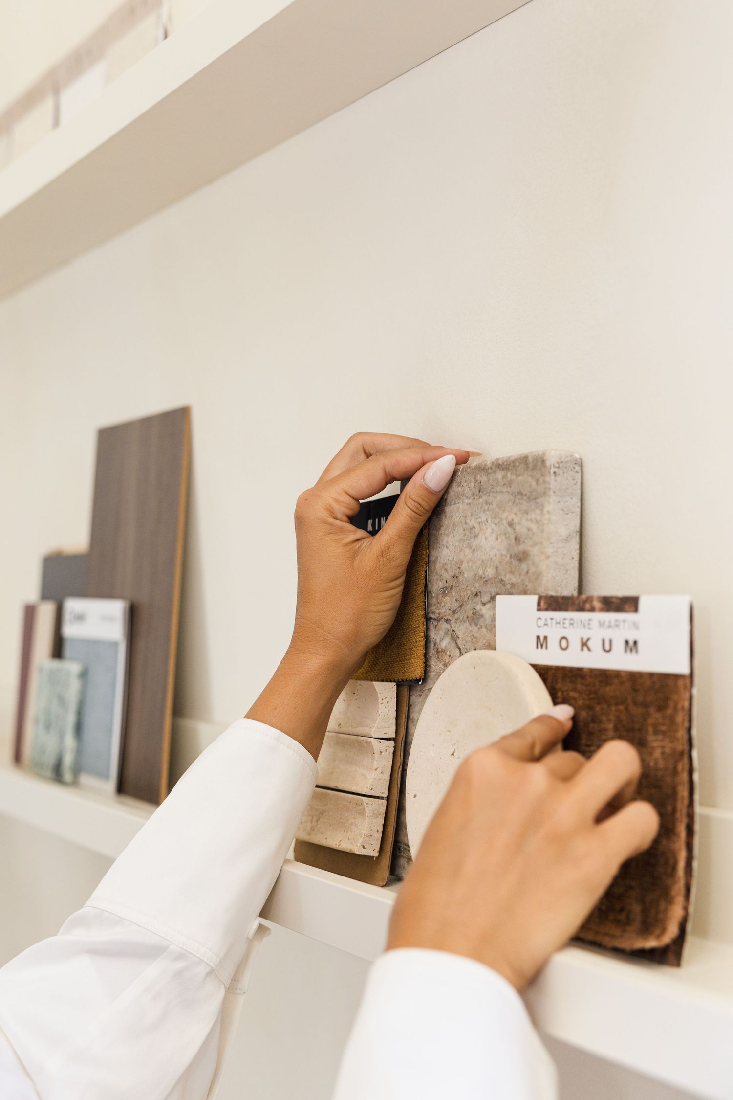 Hands selecting material and fabric samples from a display shelf in an interior design studio.