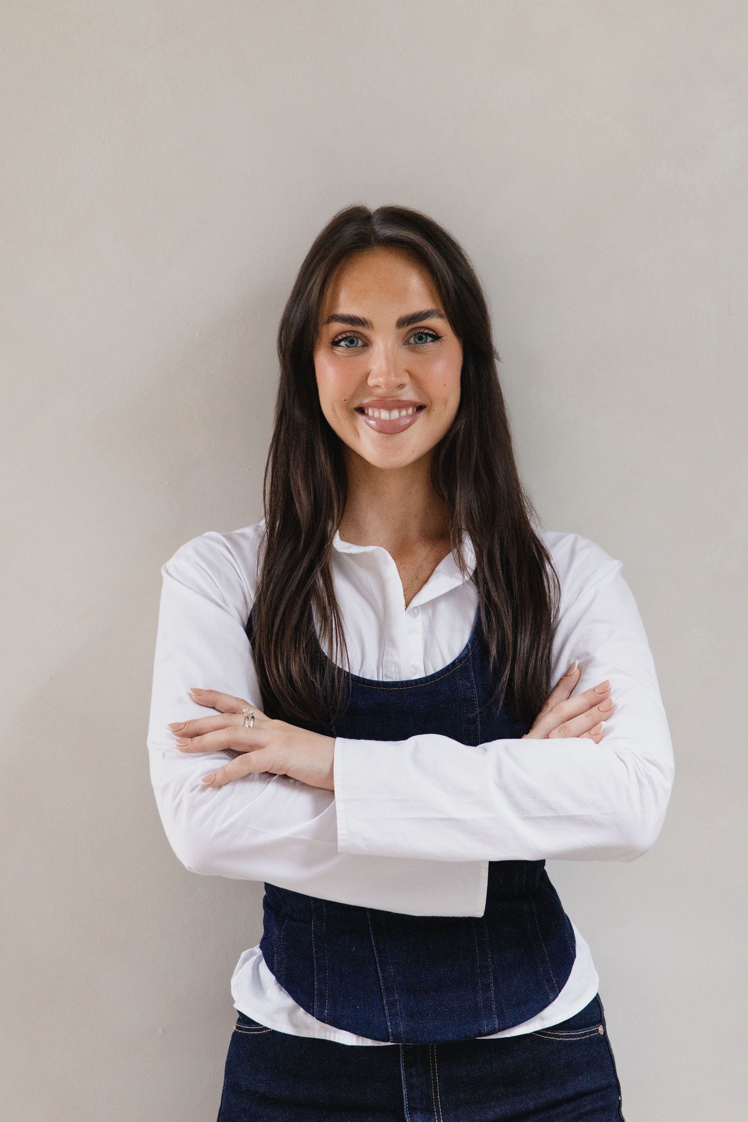 Smiling young woman with long dark hair, wearing a white shirt and denim overalls, standing with arms crossed.