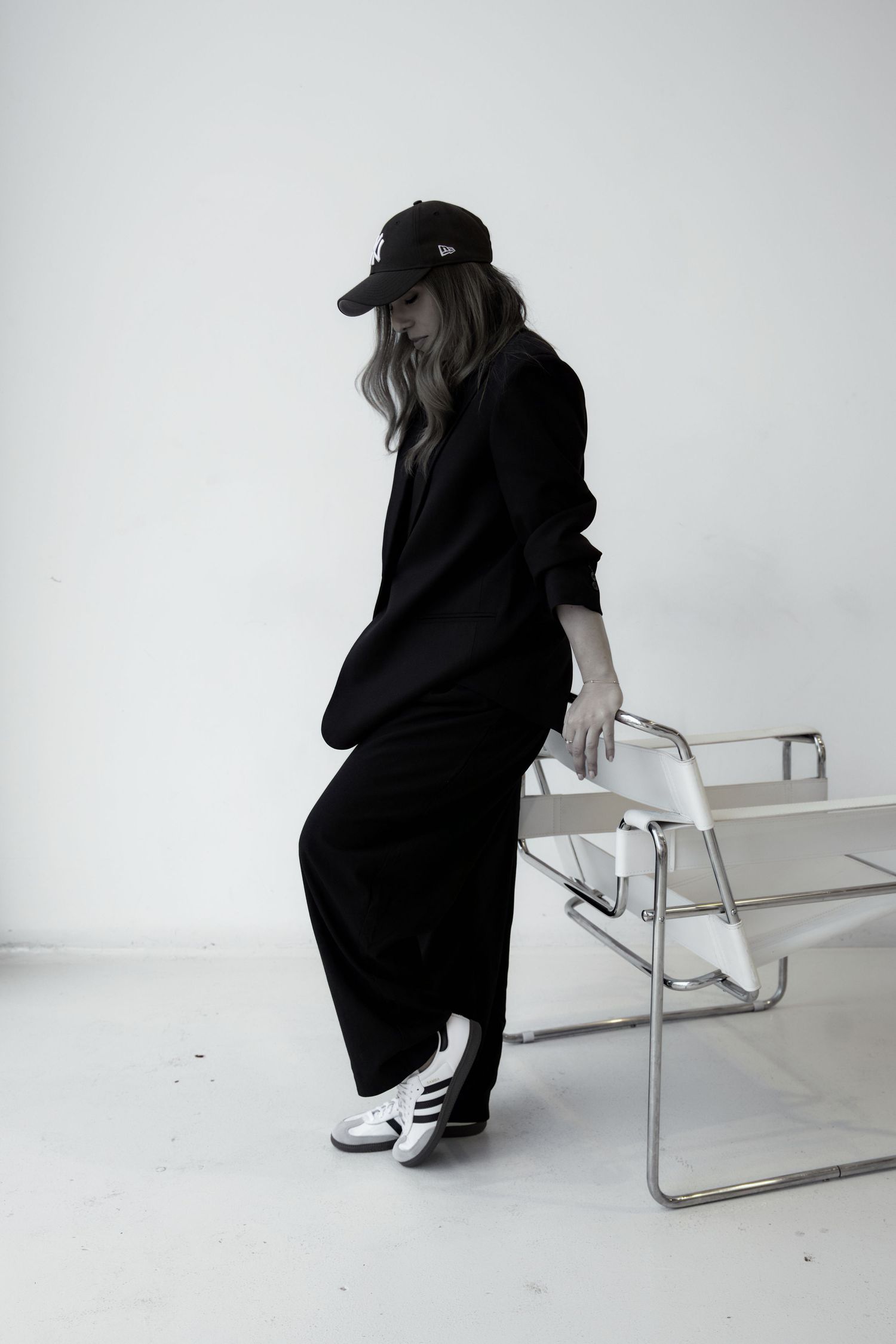 Woman in black oversized suit, cap, and Adidas sneakers leaning on a Wassily chair in a minimalist white studio.