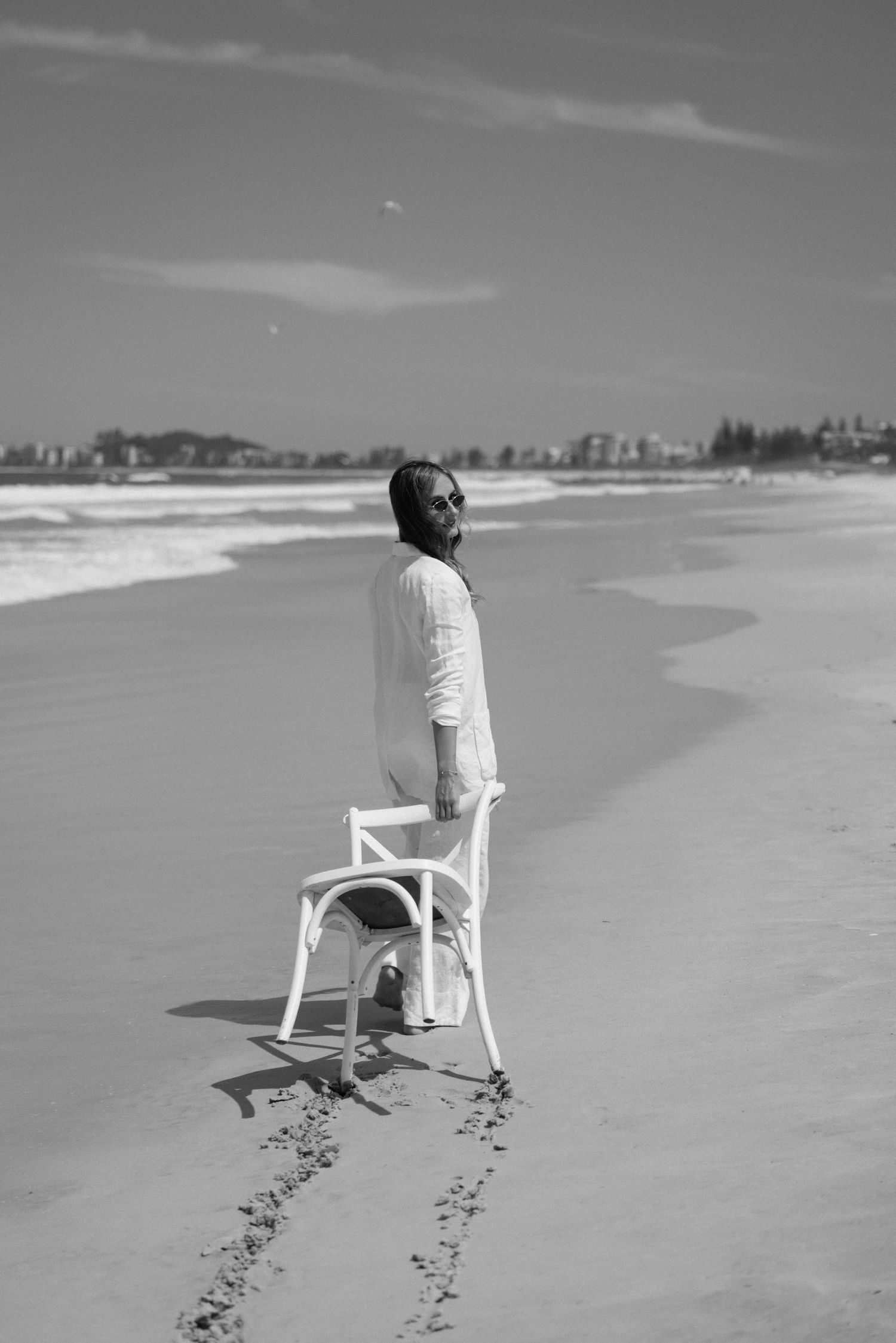 Woman in white outfit standing with a chair on a sunny beach in black and white photo.