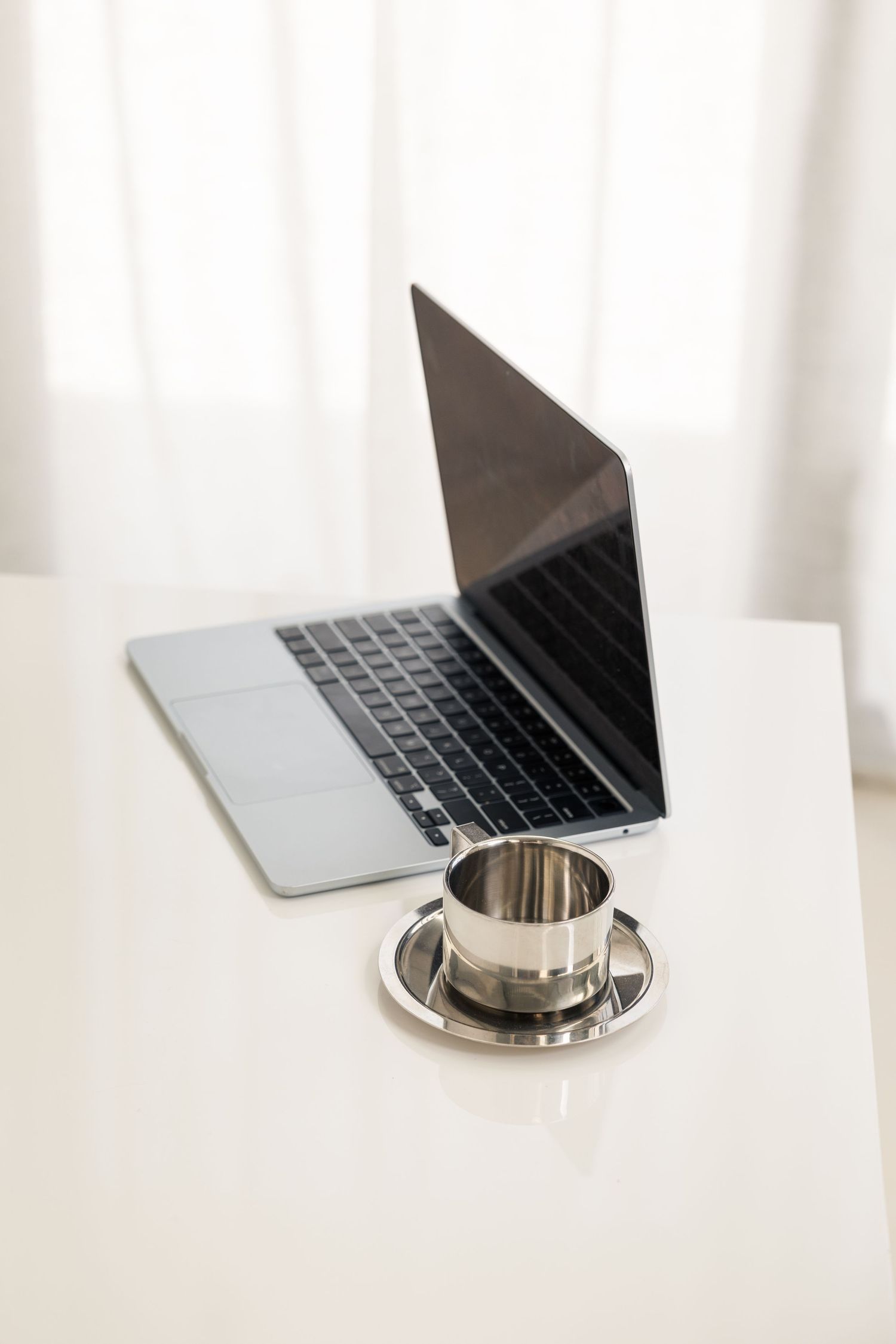 Minimalist workspace setup with silver laptop and metallic cup on white surface.