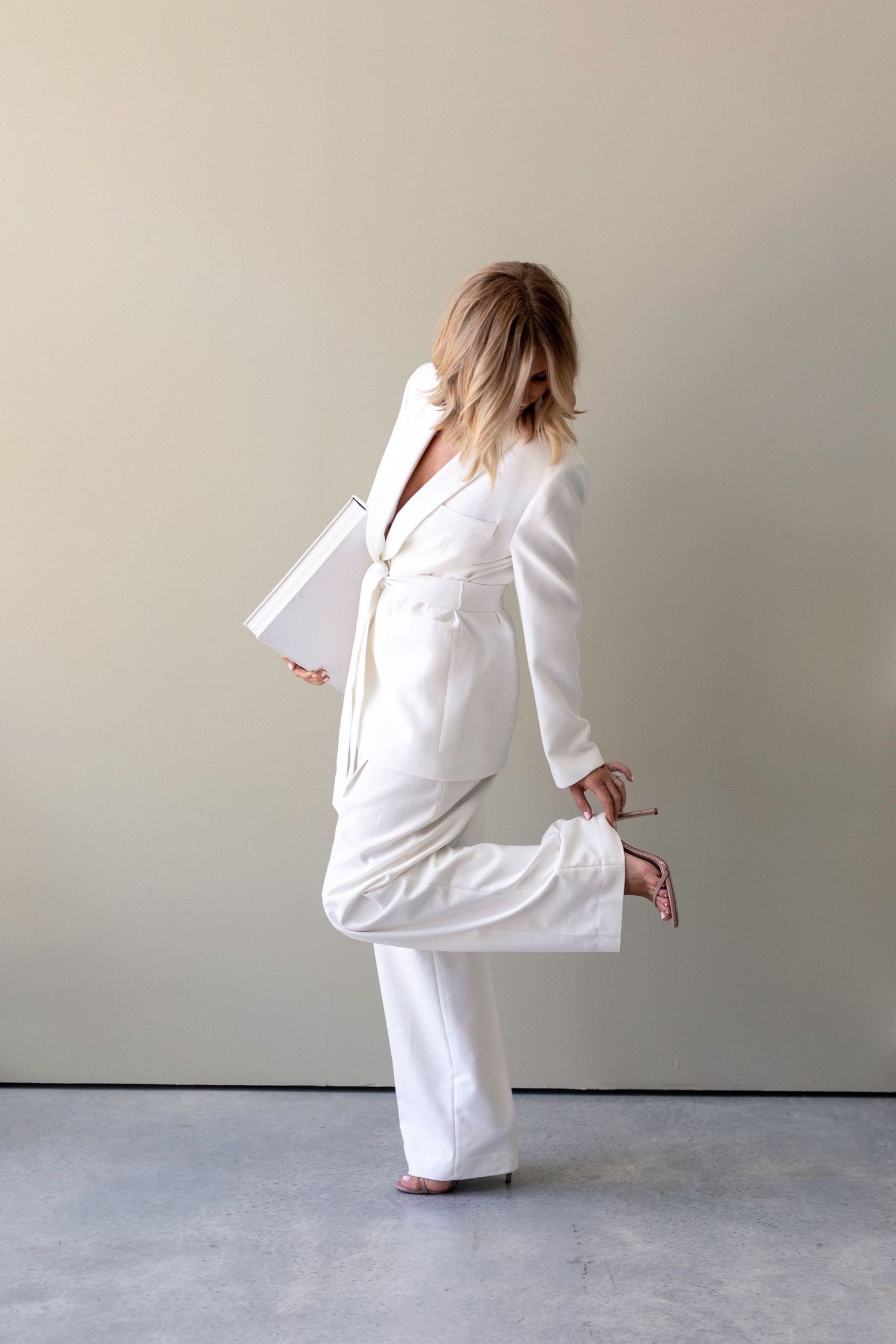 Woman in a white belted pantsuit holding a clutch, posing dynamically against a neutral wall.