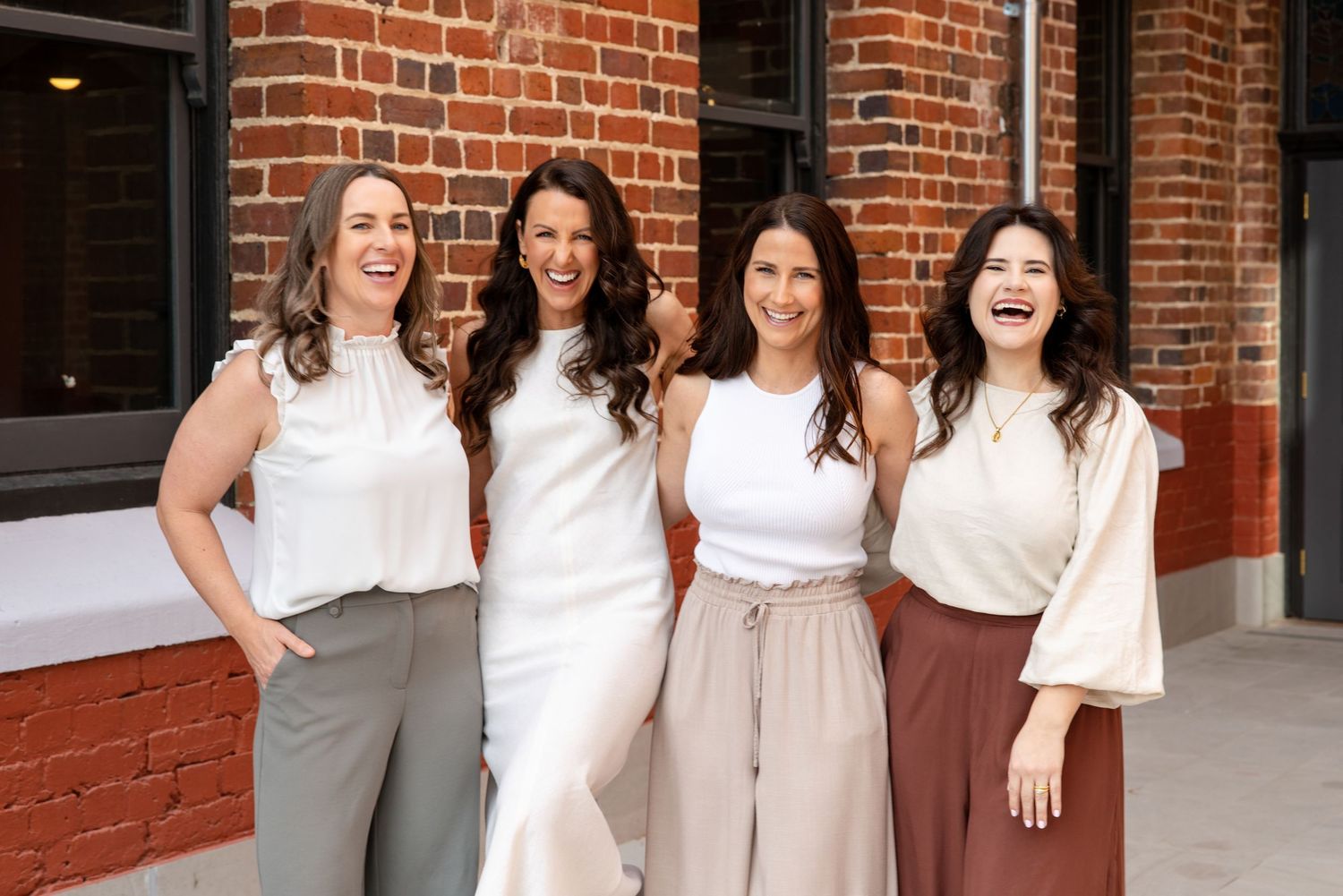 Four smiling women in neutral tones pose together in front of a brick wall outdoors.