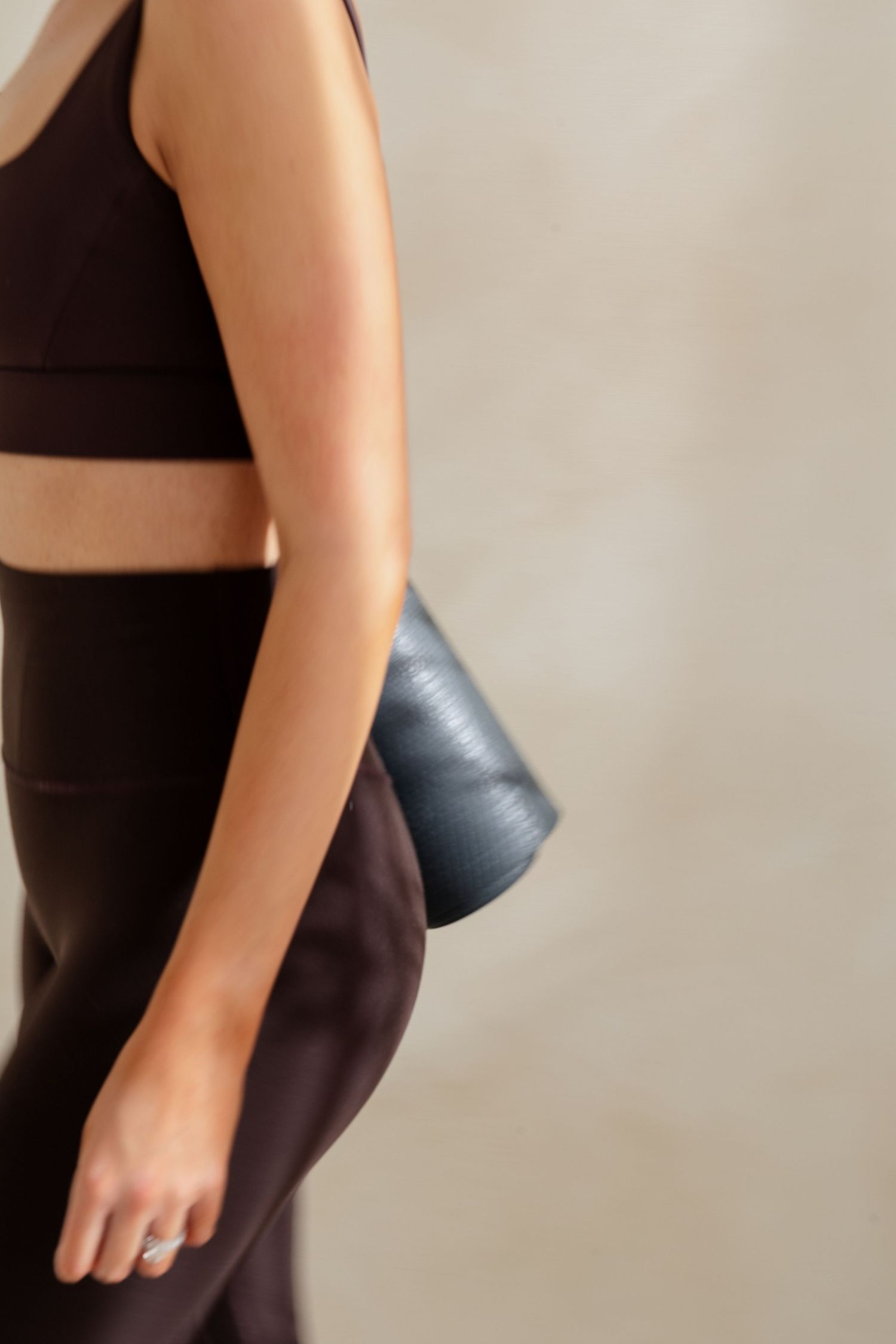 Close-up of a woman in a dark crop top and brown leggings carrying a rolled yoga mat.