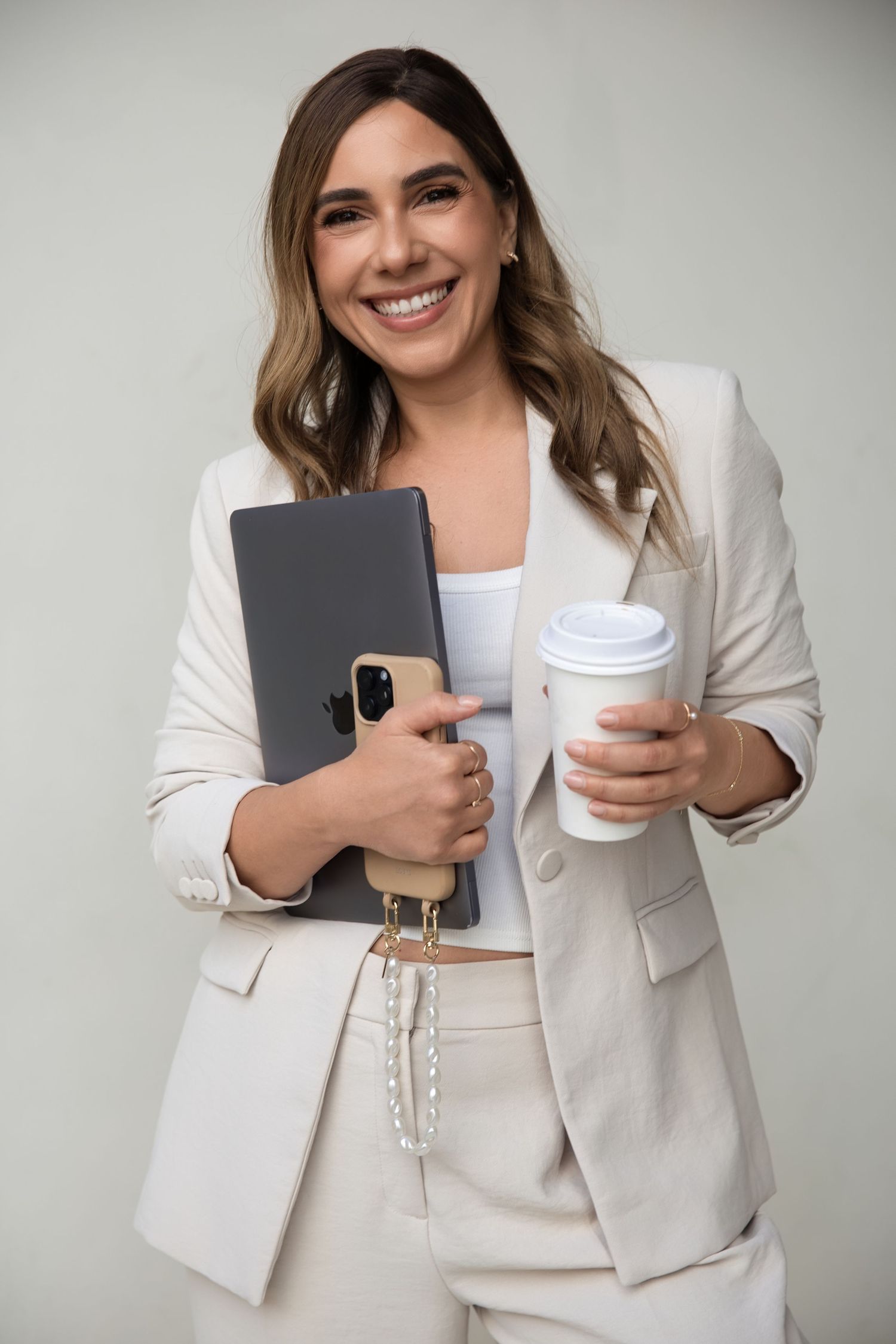 Smiling woman in cream suit holding a gray laptop, iPhone, and white takeaway coffee cup.