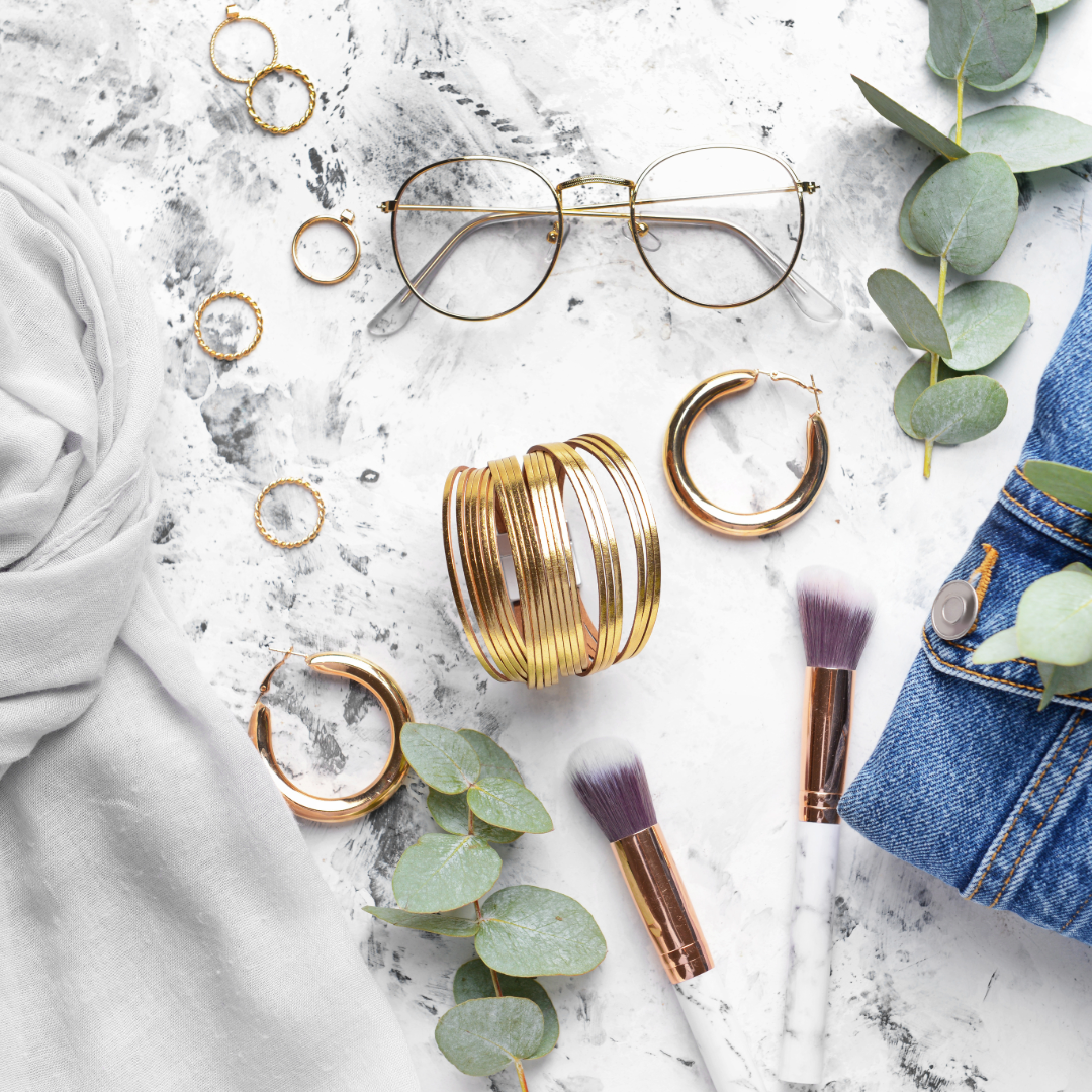 Flatlay of gold jewelry pieces including rings and bracelets on white marble surface with eucalyptus leaves and makeup brush.