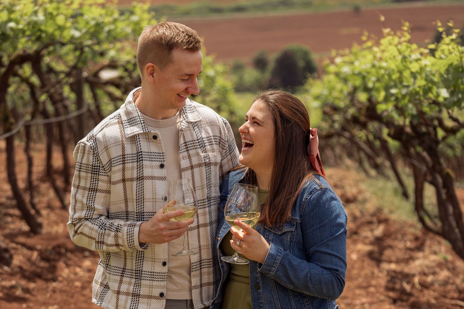 A couple shares a joyful moment while holding mugs in a scenic vineyard setting during autumn.