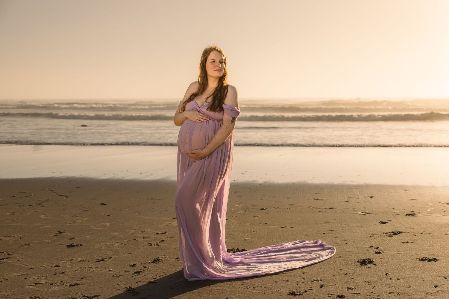 Expectant parent in flowing lavender gown poses elegantly on beach at sunset with train flowing in breeze.