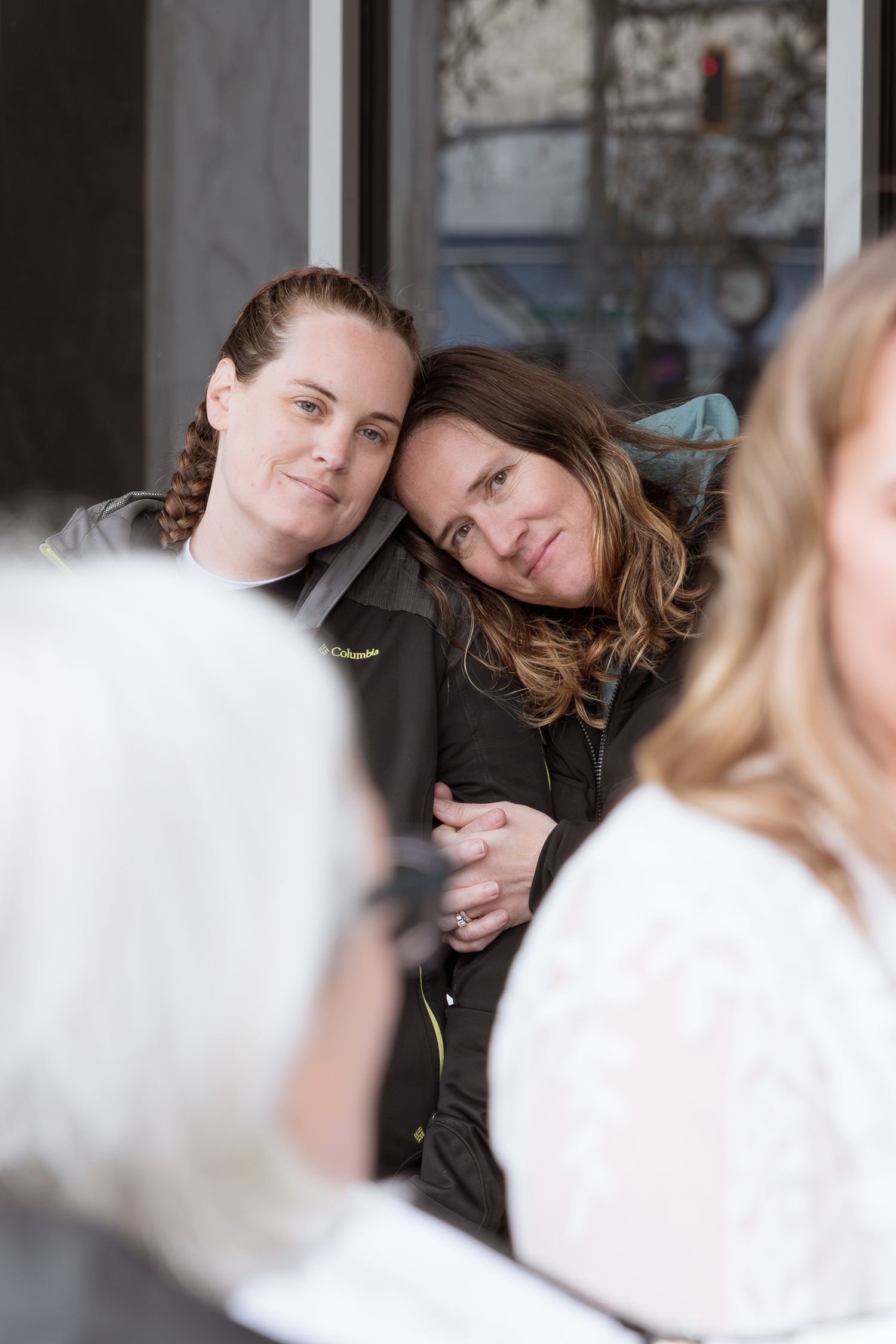 Two women leaning heads together smiling warmly, sharing an affectionate moment outdoors near a building entrance.