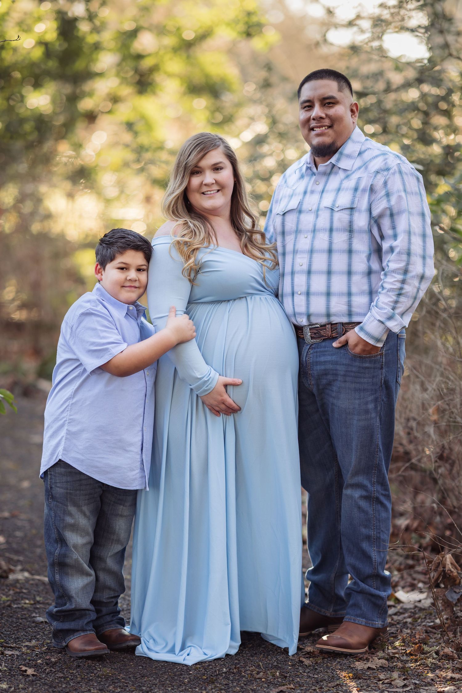 Family maternity photo outdoors showing expectant mother in light blue dress with two family members on a nature path.