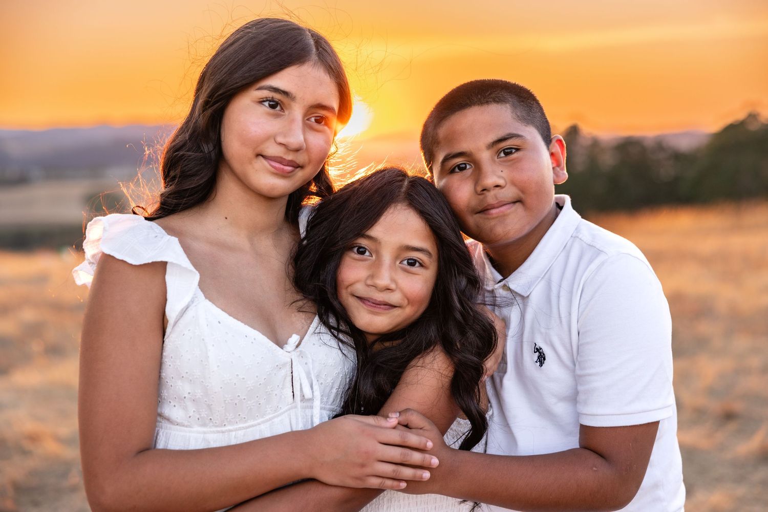 Three siblings pose together at golden hour in a field, wearing white outfits, smiling warmly at the camera.
