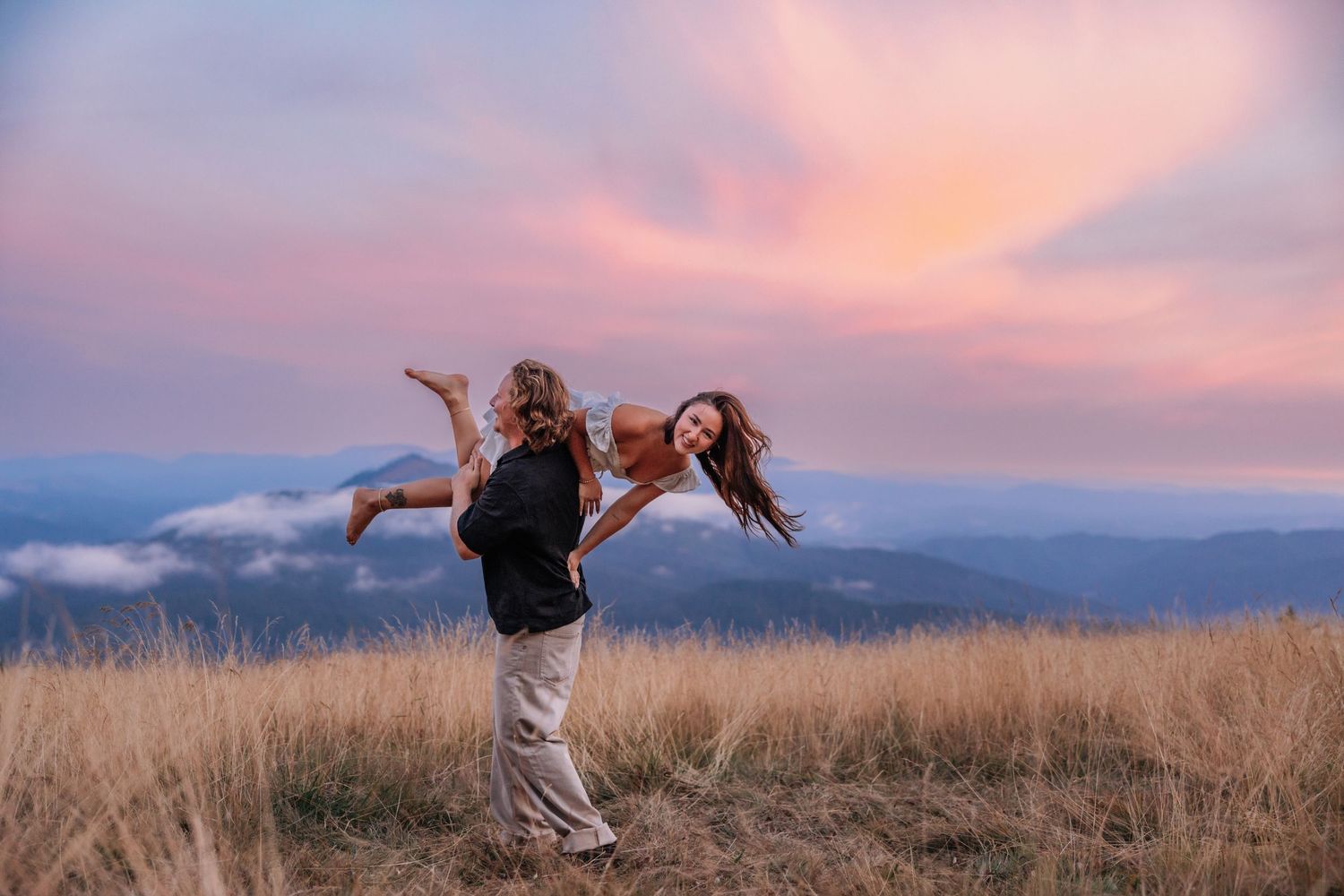 Couple dancing joyfully in golden meadow with stunning pink sunset sky and mountain landscape backdrop.