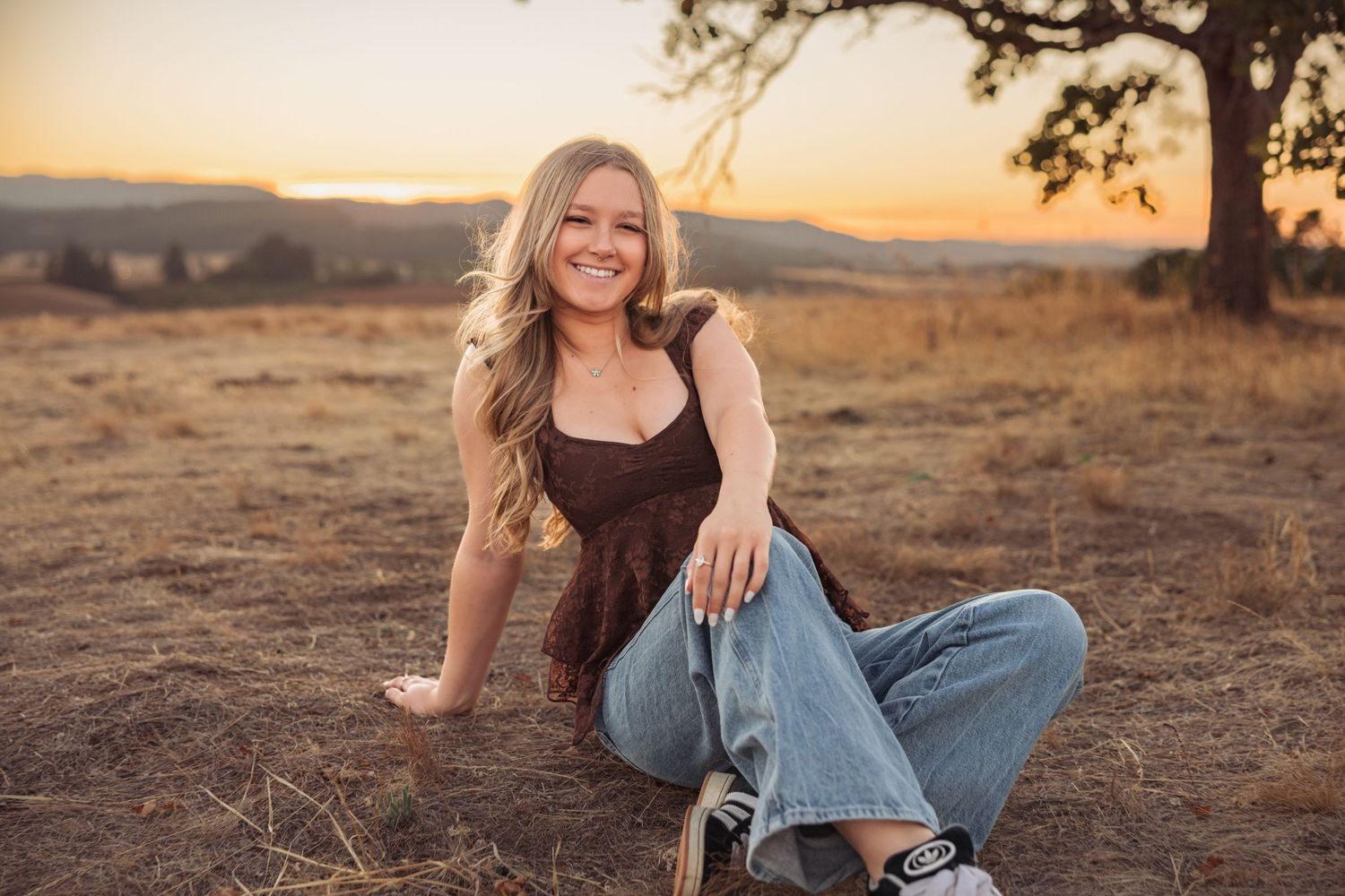 A person in casual denim sits on a grassy hillside during golden hour as warm sunlight illuminates the scenic landscape.