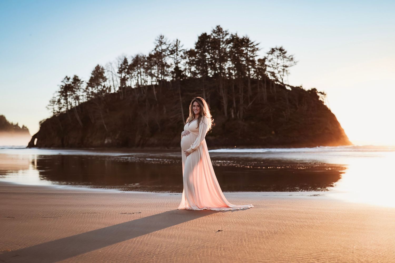 Woman in a flowing pink dress stands on beach at sunset during a photo session with Salem, Oregon maternity photographer.