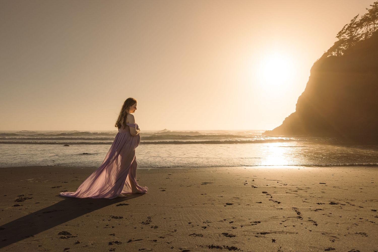 A figure in flowing pink dress walks on sandy beach at golden sunset with cliffs in the background.