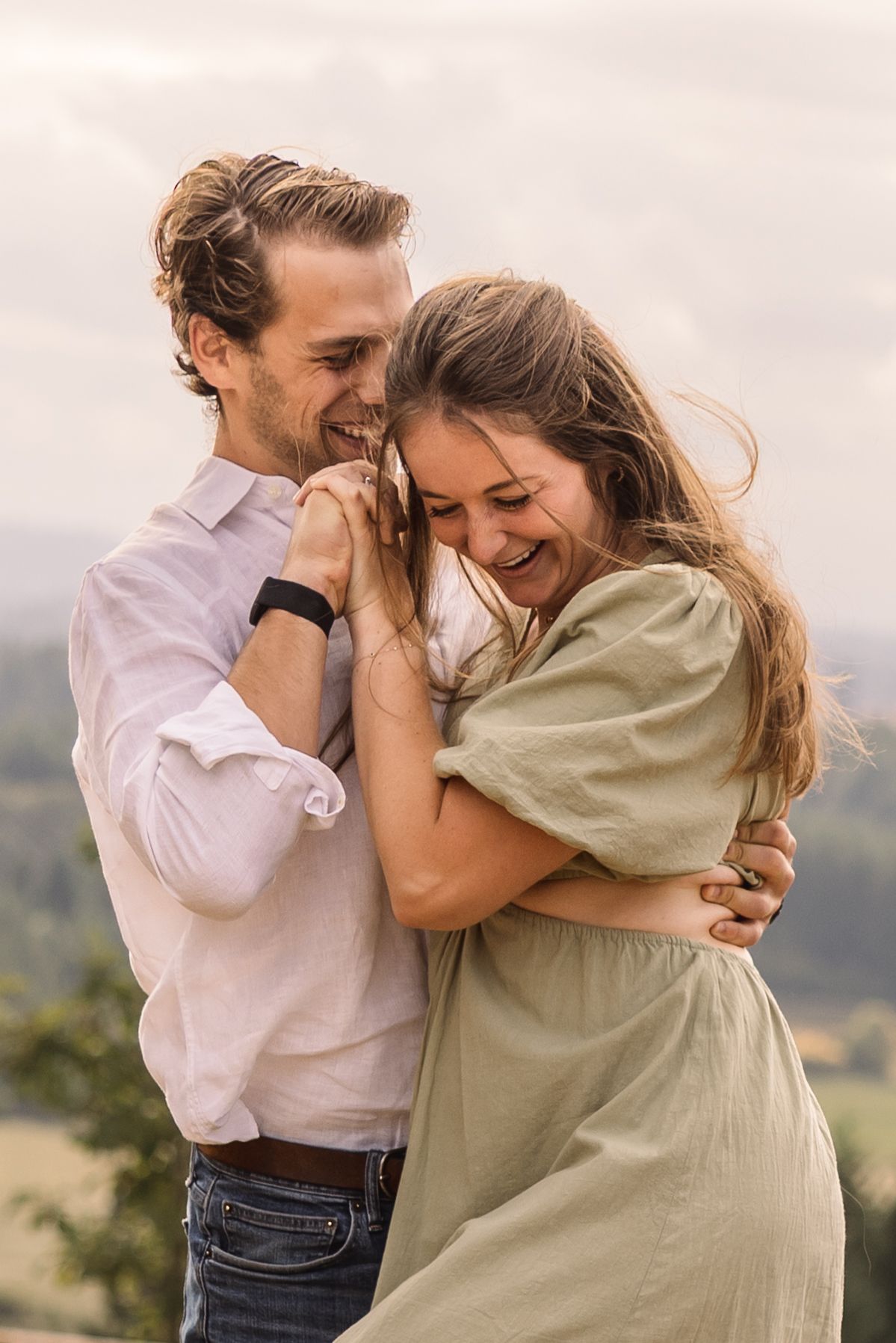 A romantic couple embraces and laughs together during a mountaintop photoshoot at sunset.