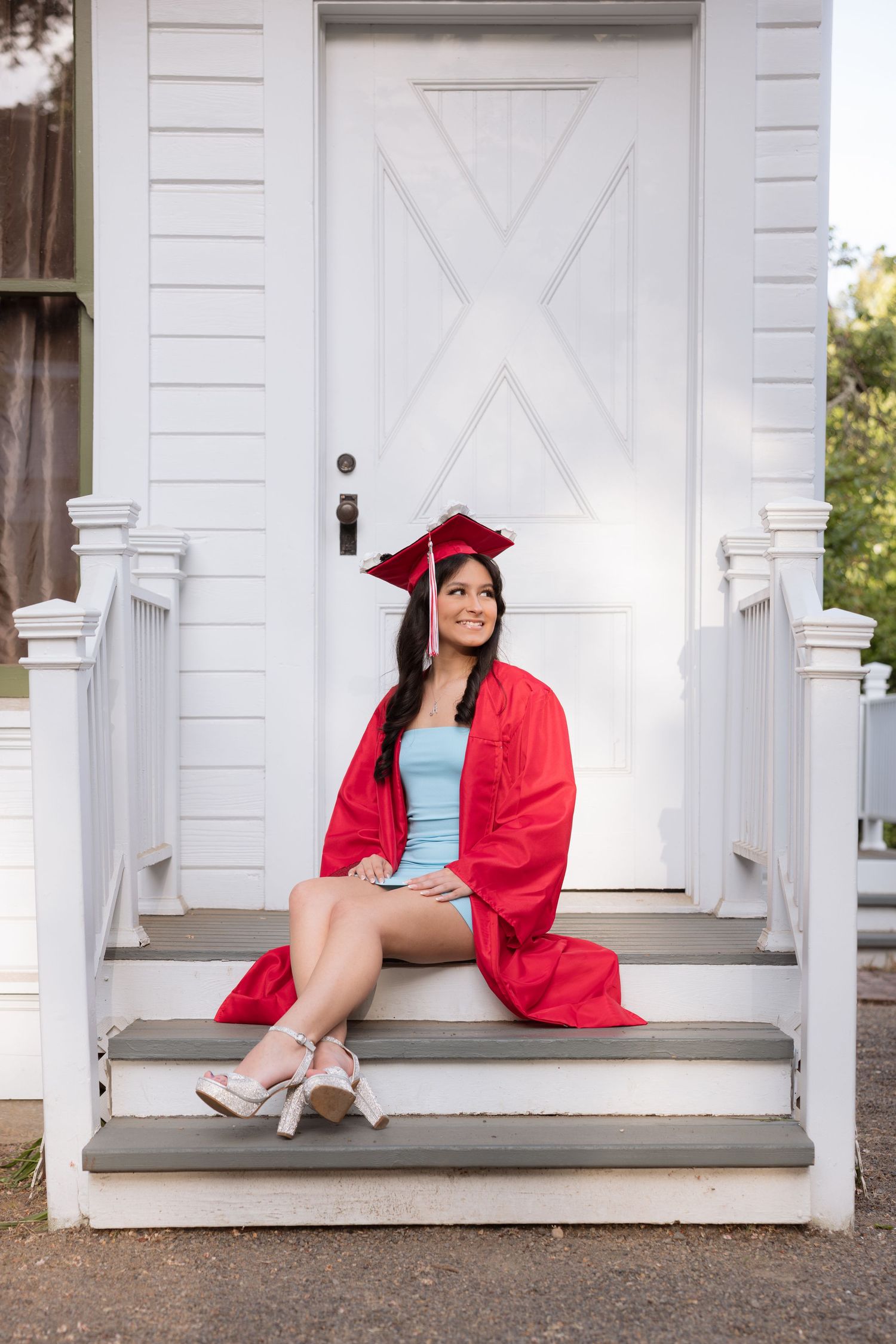 Young woman in red graduation cap and gown sitting on white porch steps, smiling and looking up.