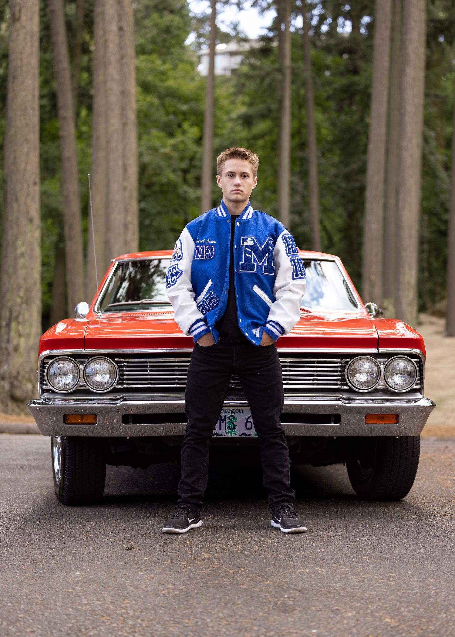 Young man in blue varsity jacket posing in front of a classic red car on a forest road.