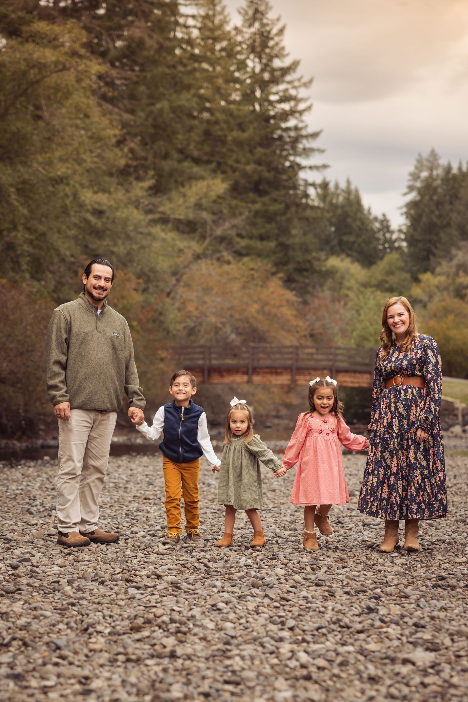 Family group stands together on rocky riverbank surrounded by autumn trees in Pacific Northwest setting.