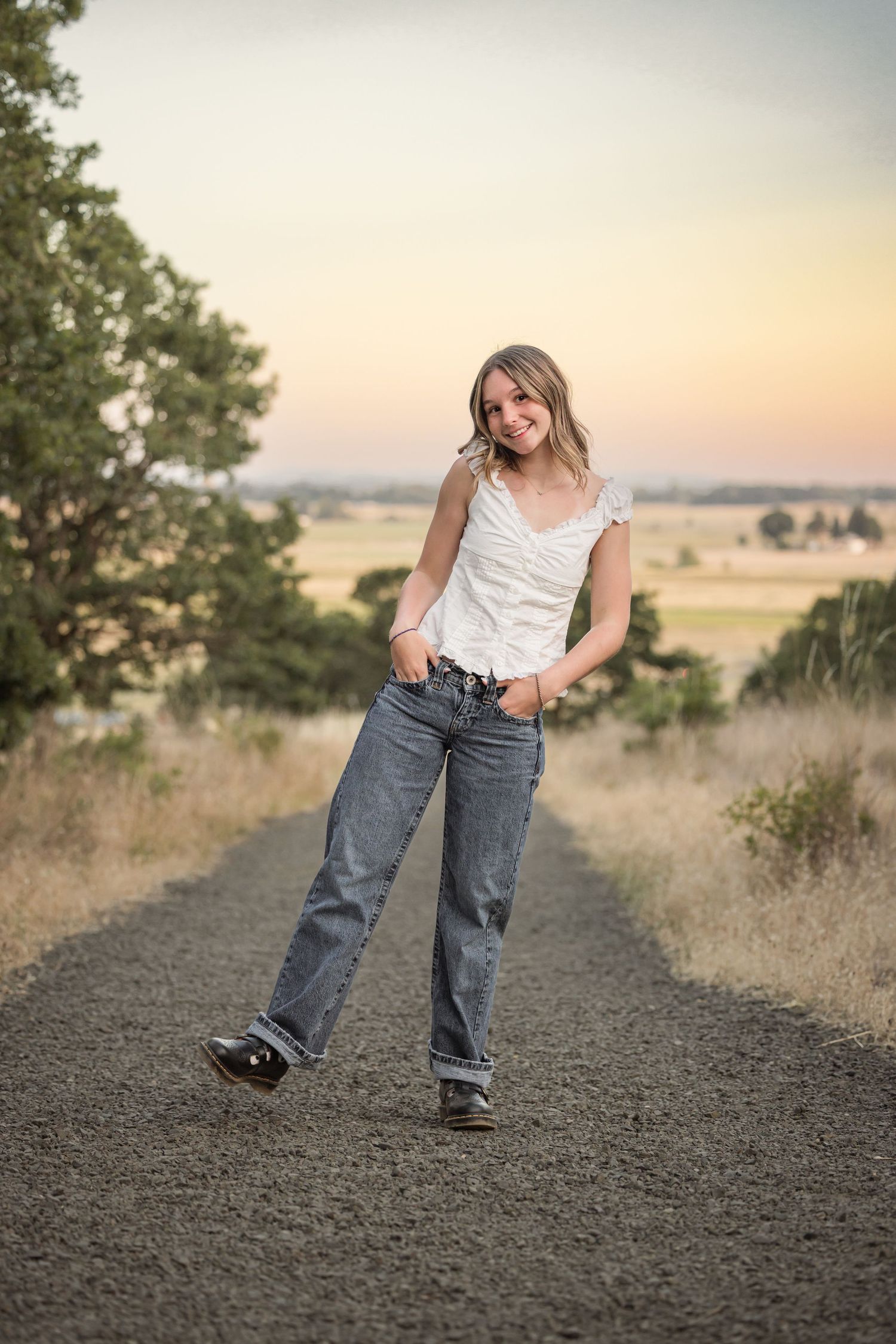 A person in casual attire poses on a dirt path at sunset with rolling hills and trees in the background.