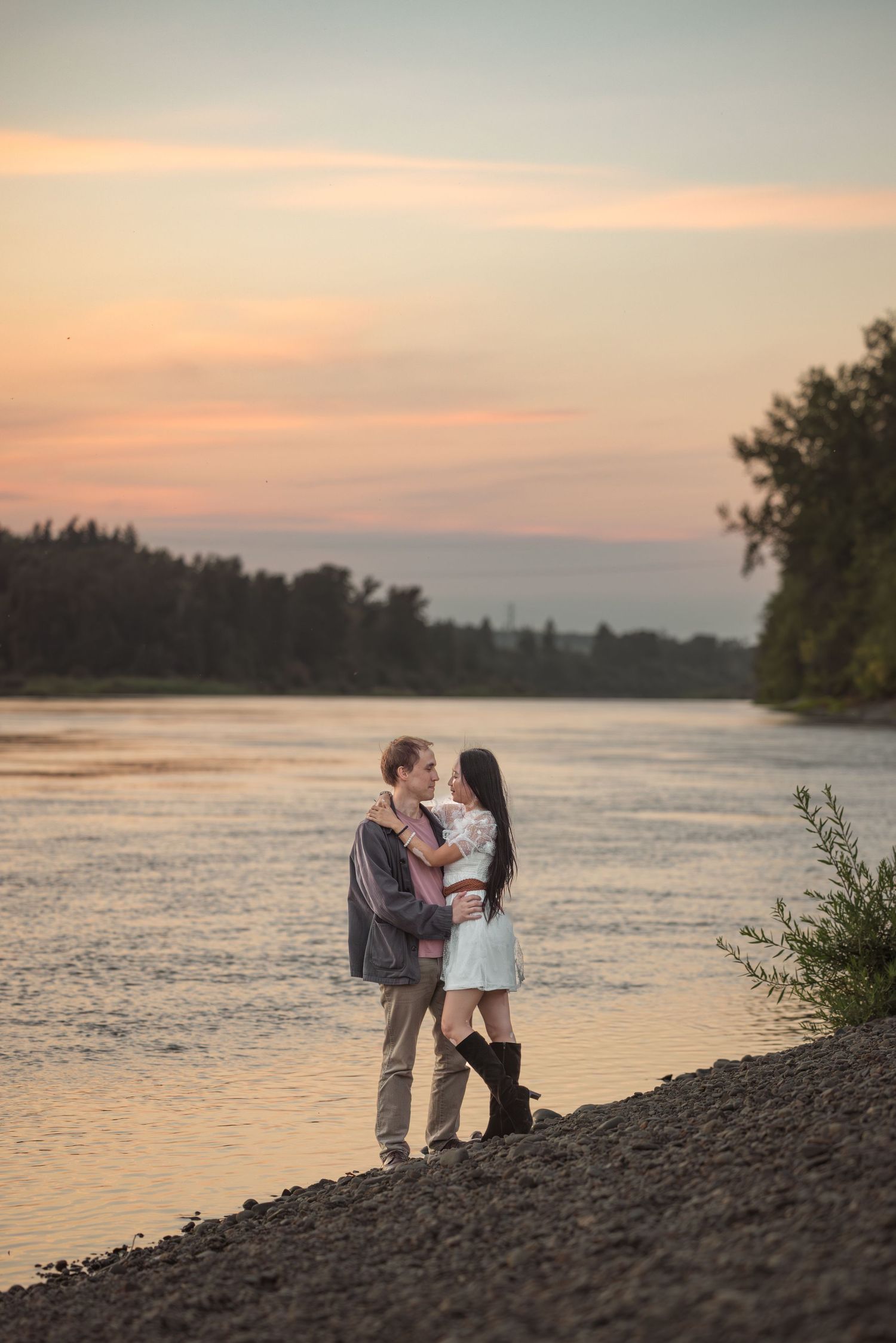 Romantic couple on riverbank at sunset with colorful pink and orange sky behind them in Salem, Oregon. Oregon engagement photographer.