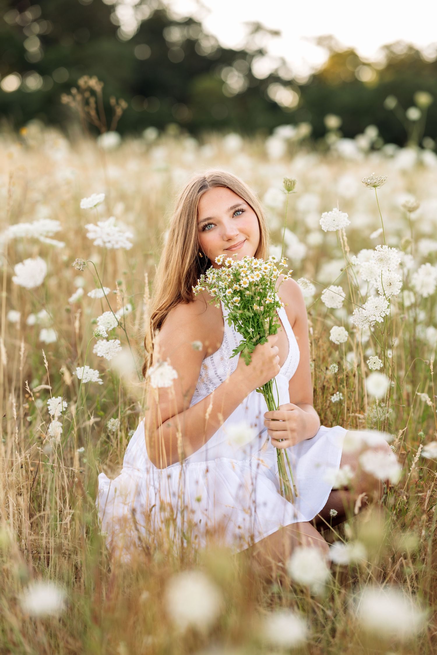 Young woman in white dress sitting in wildflower field holding bouquet of white flowers, smiling in golden sunlight.