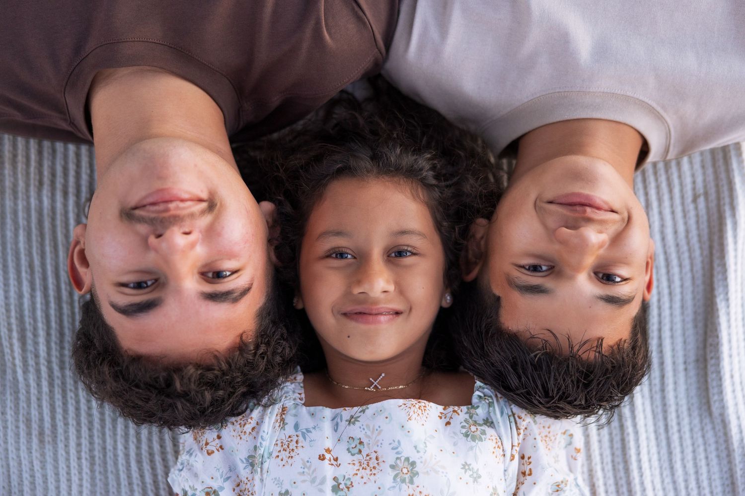 Three smiling family members lying on a bed looking up at the camera, two adults and a young girl in the middle.
