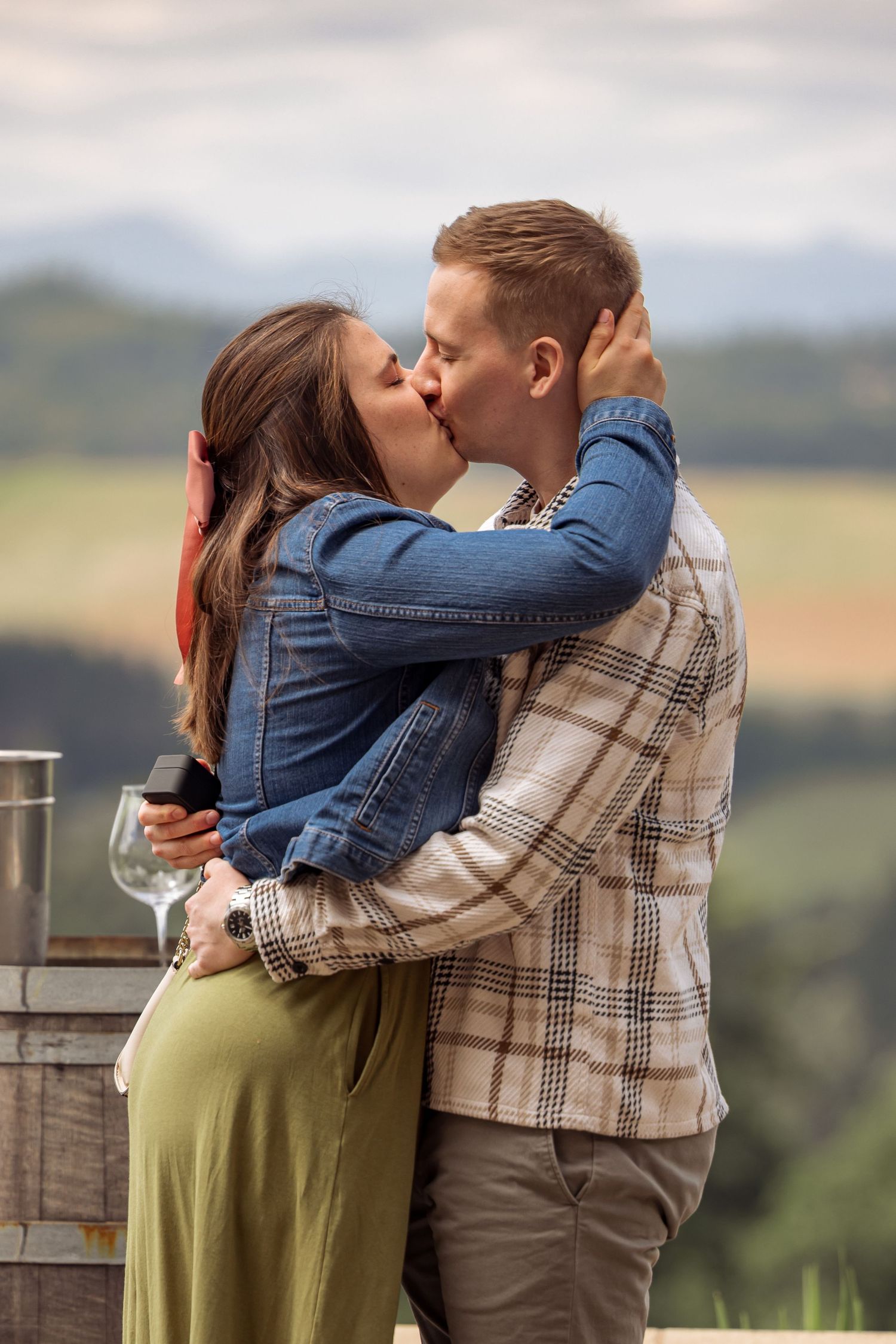 A couple embraces in a passionate kiss against a backdrop of rolling hills at a scenic winery overlook.
