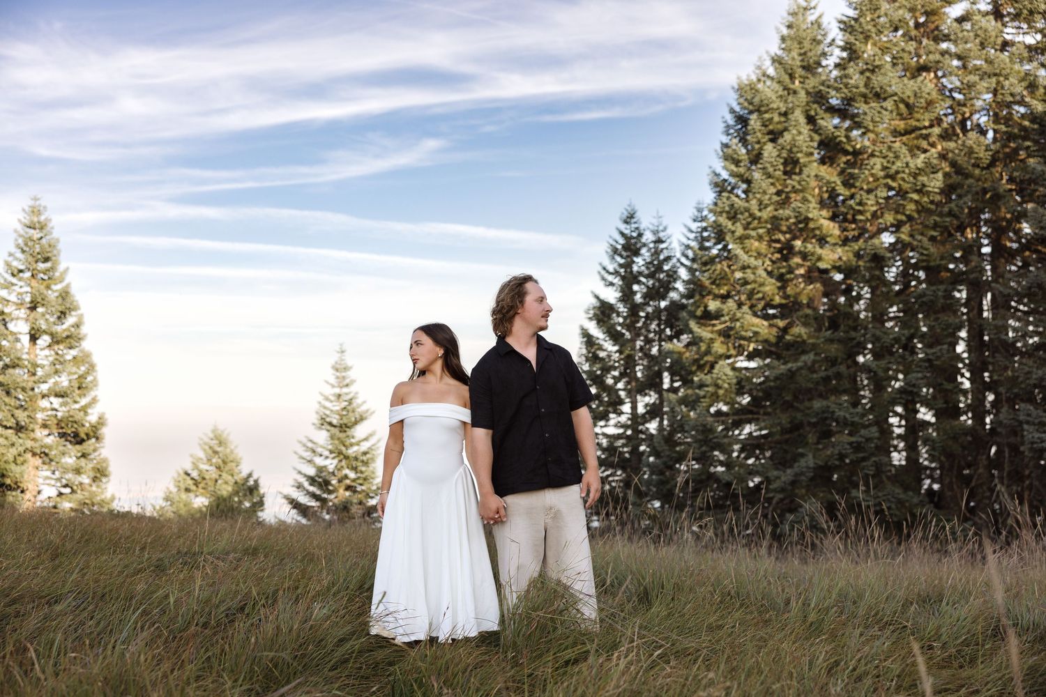 Couple standing in meadow with evergreen trees, woman in white dress, man in black shirt, golden hour sky.
