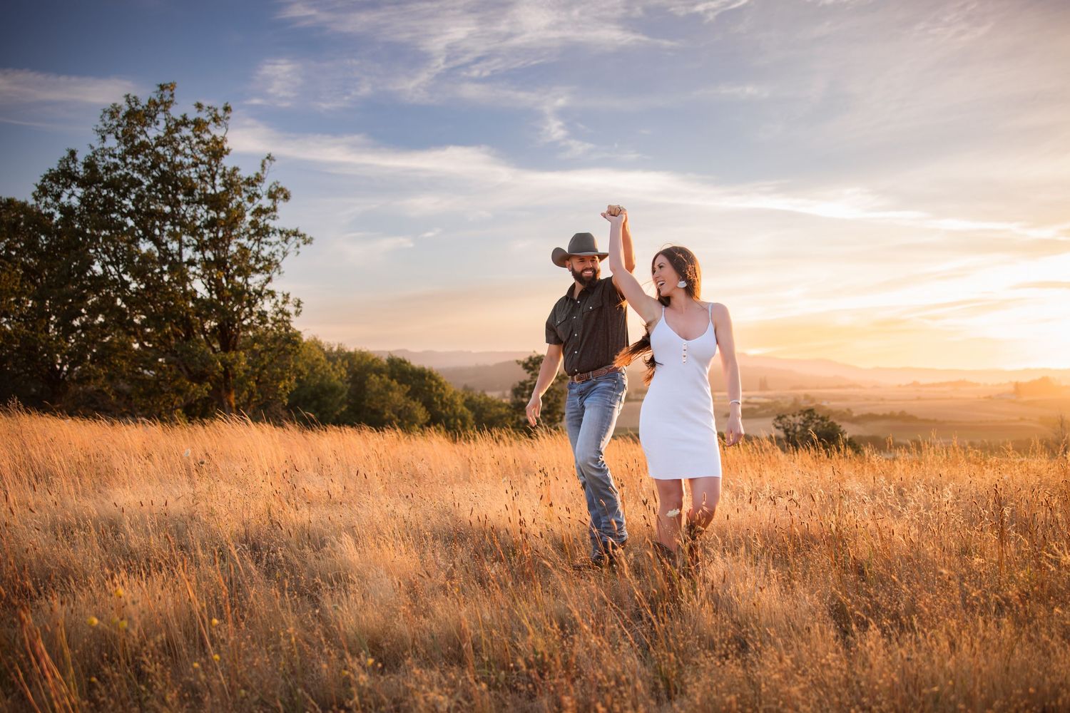 Couple dancing in golden field at sunset, man in cowboy hat and woman in white dress, romantic outdoor engagement photo.