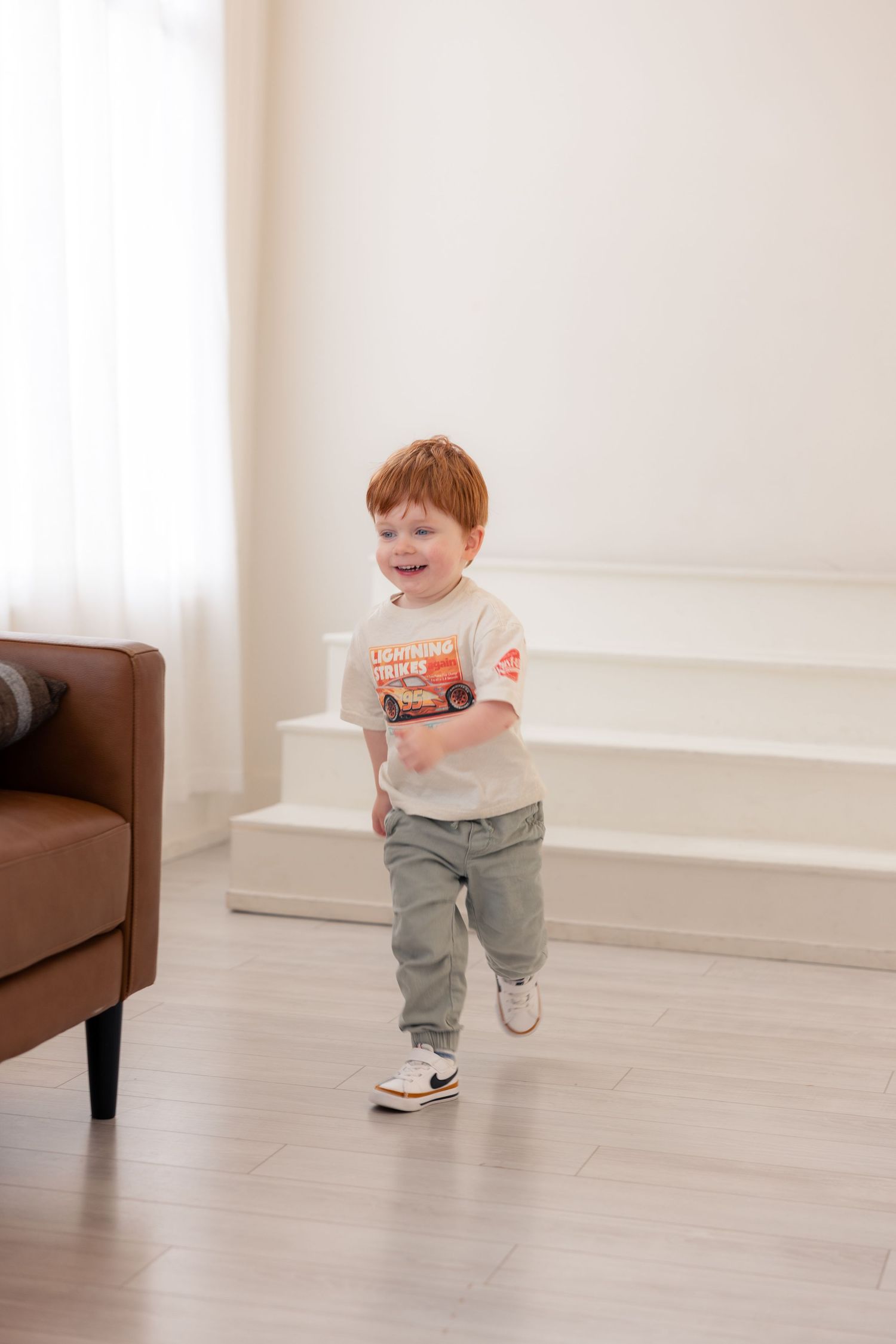 Toddler in gray pants and white t-shirt running and playing excitedly on light hardwood floors next to brown furniture.
